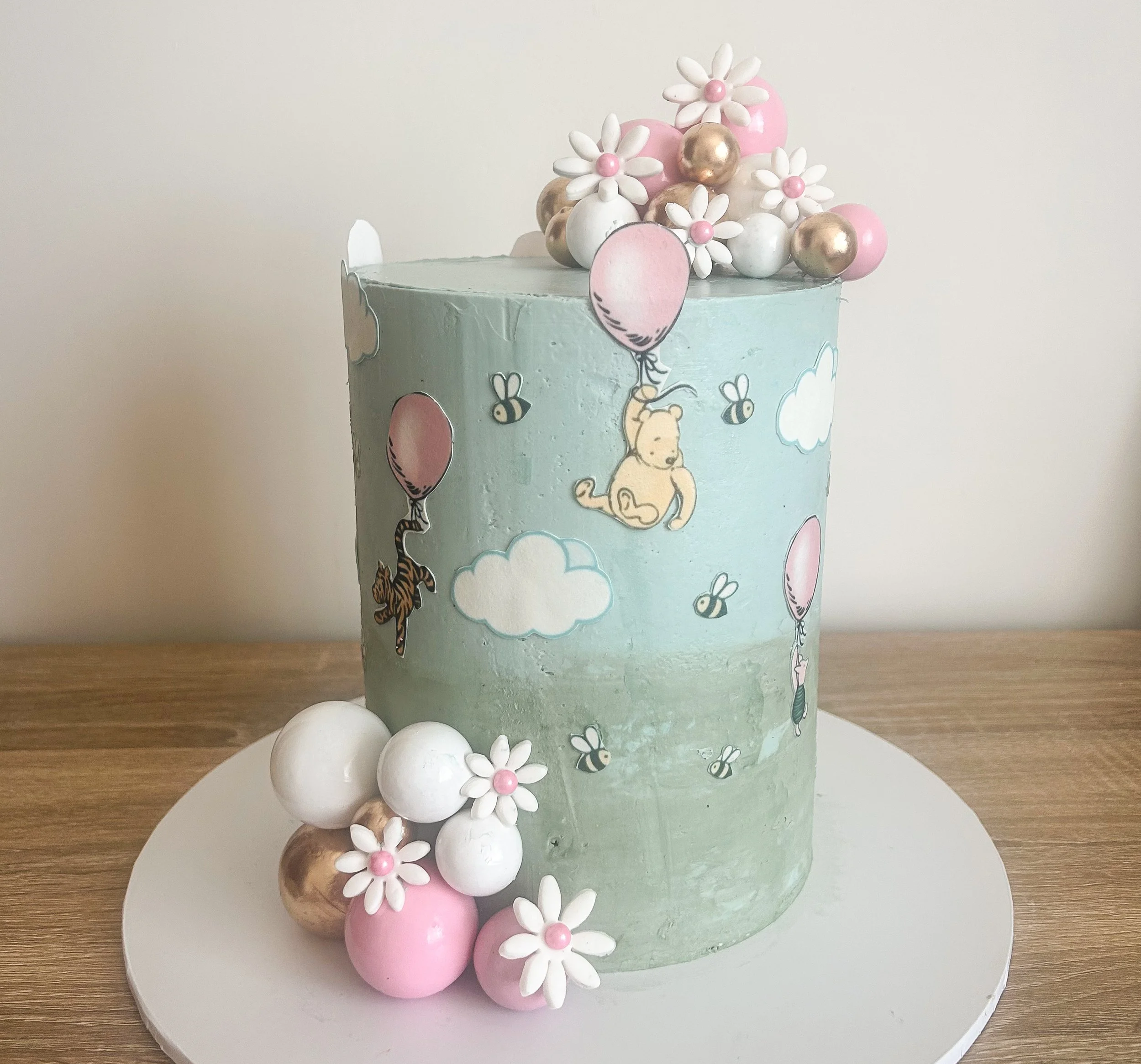 Light blue birthday cake decorated with Disney Winnie the Pooh theme featuring clouds, bees, balloons, and honey pots. Topped with white, pink, and gold spherical ornaments and white daisy-shaped flowers with pink centers.