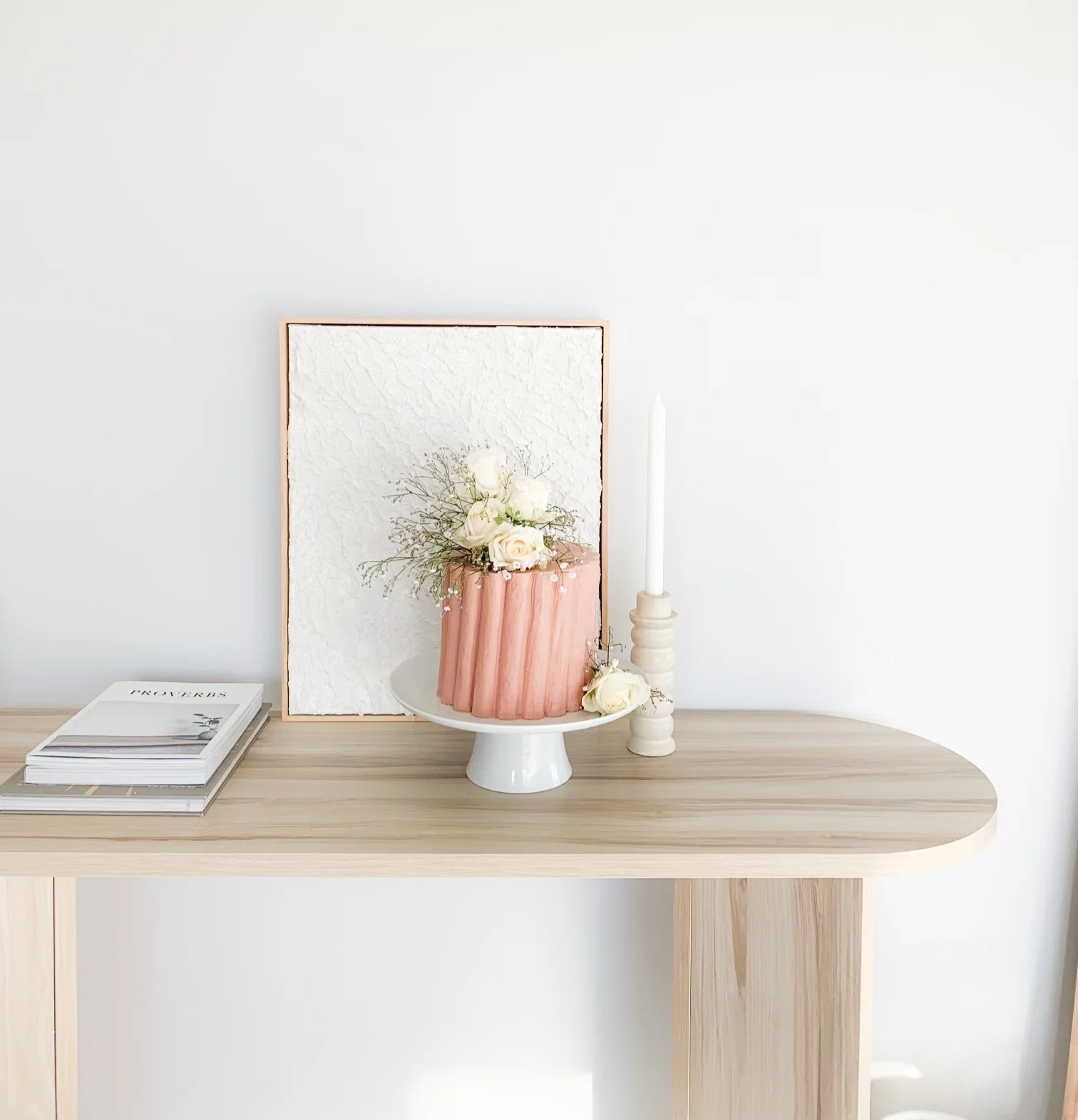Decorative table with a pink cake topped with white flowers and greenery, a framed textured artwork, a tall white candle on a beige candlestick, and a stack of books on a light wood table