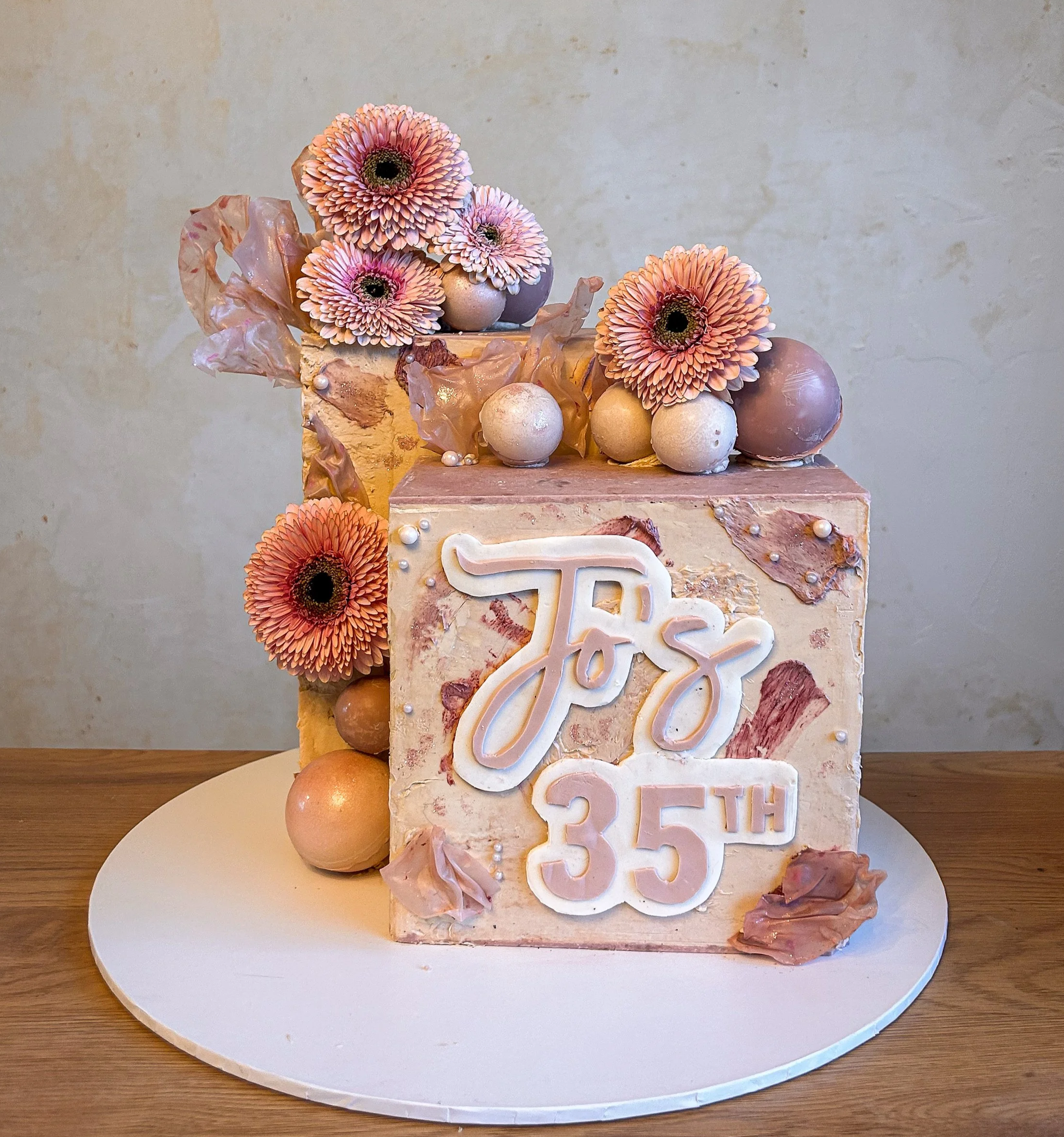 A birthday cake decorated with pink gerbera daisies, gold and pink spheres, and edible leaves. The front of the cake has a white and pink sign that reads 'Jo's 35th'.
