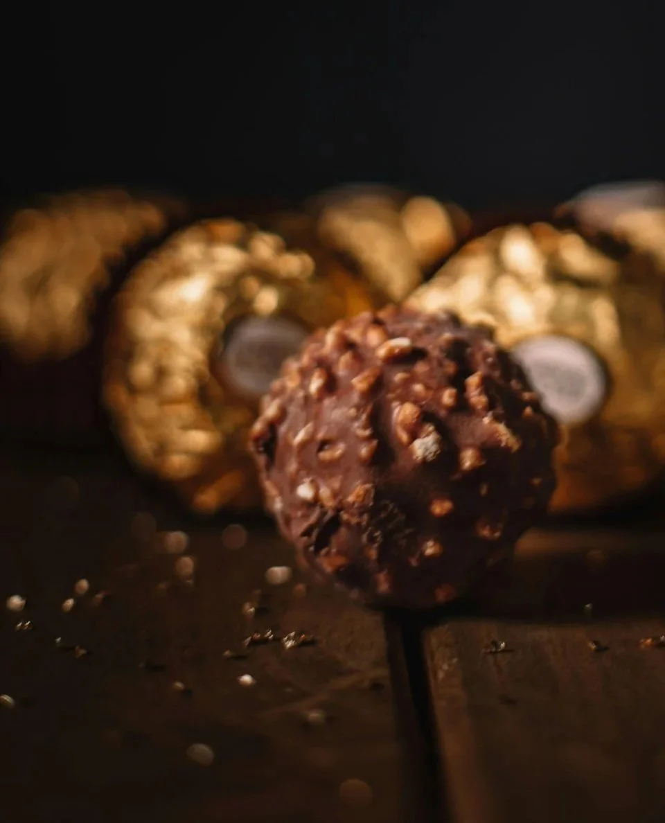 Close-up of a chocolate truffle on a skewer in front of a platter of assorted chocolates and gold-wrapped candies.