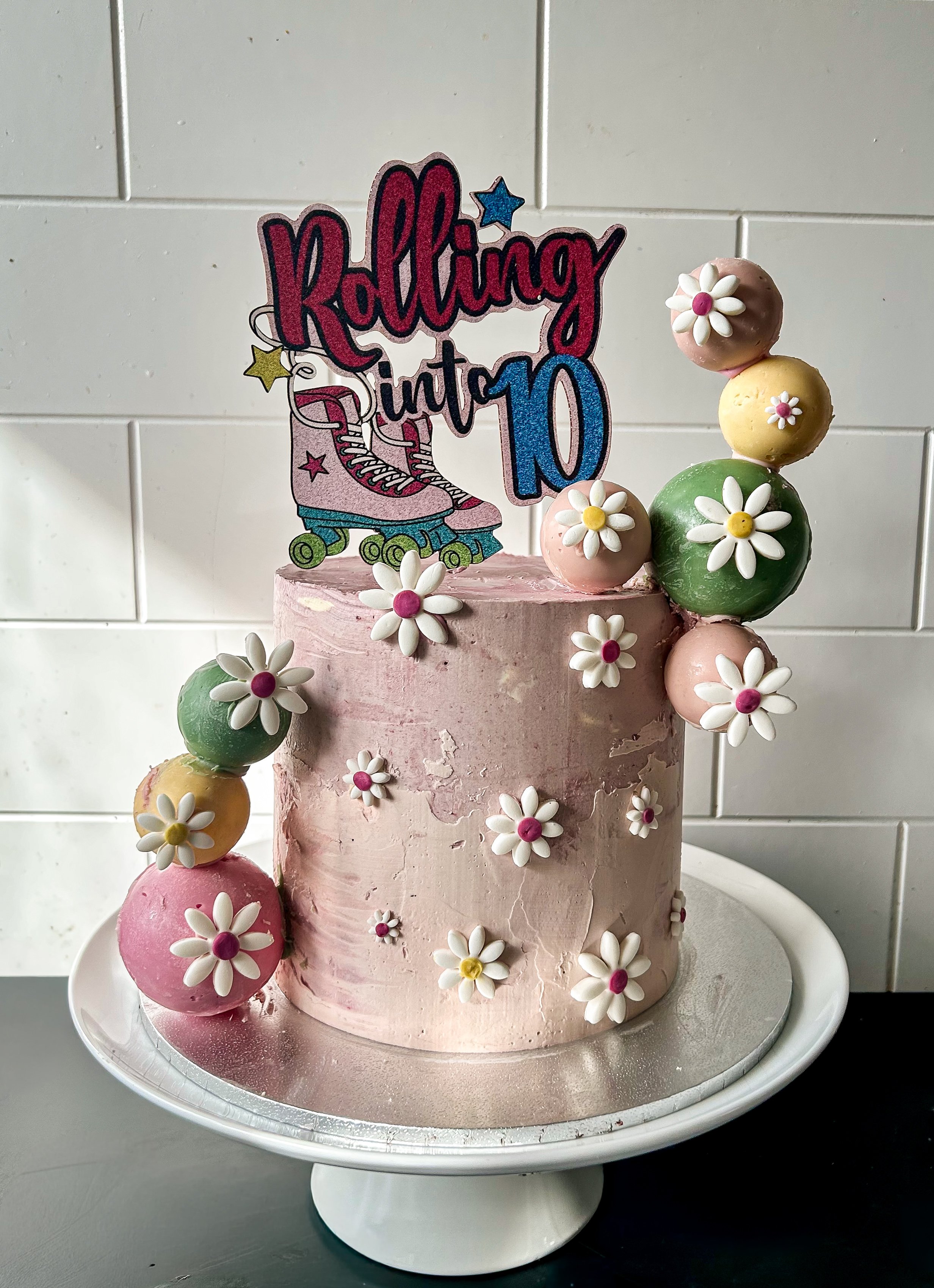 A pink birthday cake decorated with white flower candies, with a topper that reads "Rolling into 10" featuring roller skates, set against a white tiled wall.