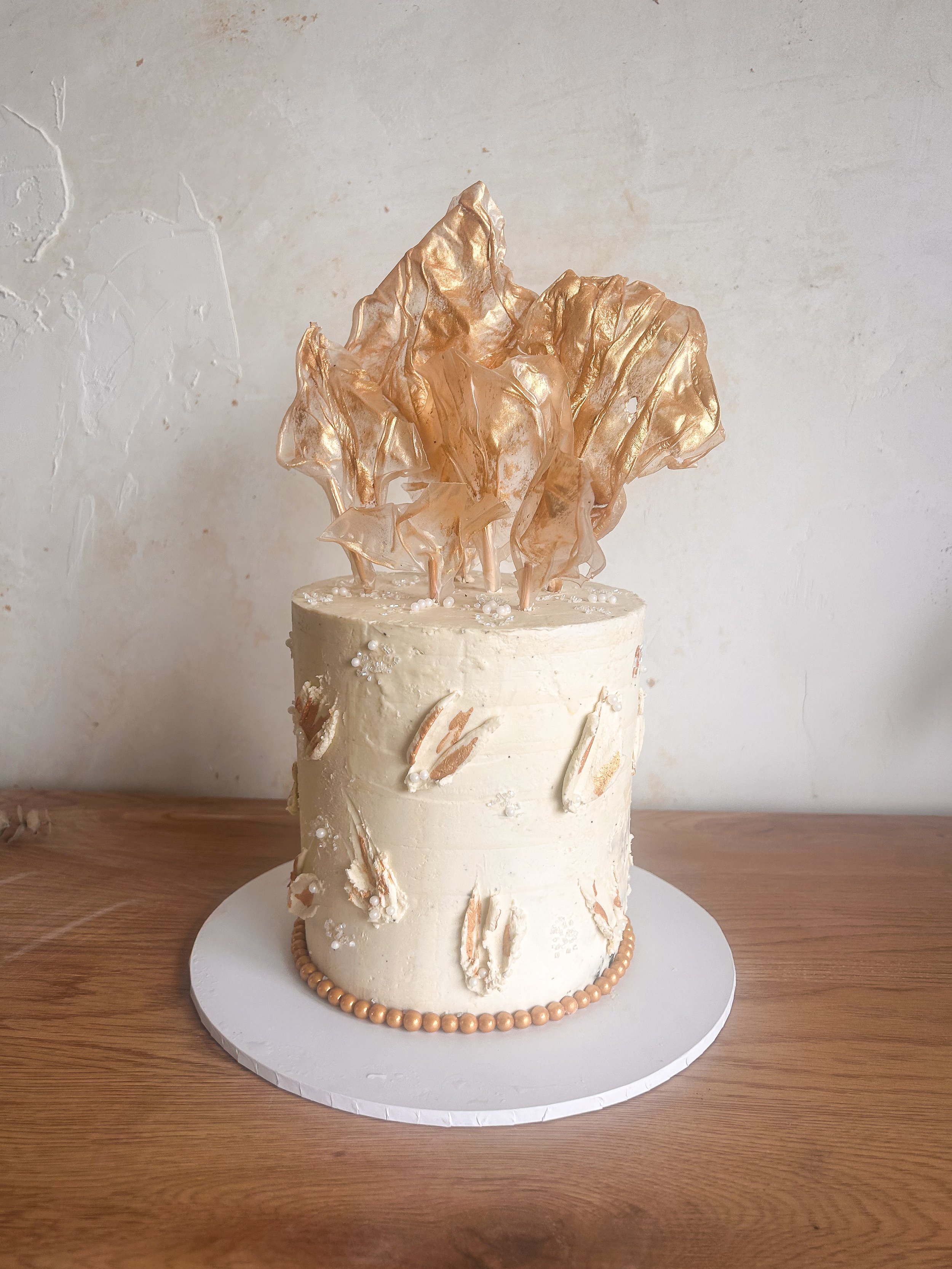 A tall, white frosted cake decorated with gold accents and a large, gold, abstract leaf topper. The cake sits on a white round base on a wooden surface.