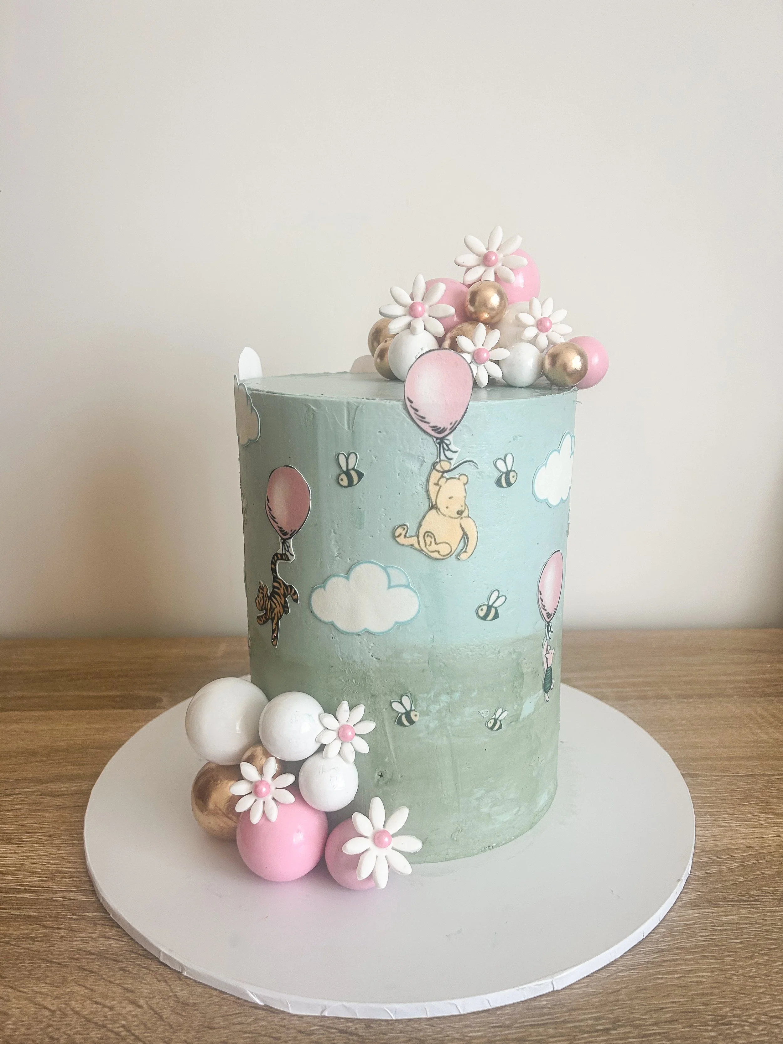 A tall birthday cake decorated with images of teddy bears, bees, clouds, and balloons, topped with pink, white, and gold spherical decorations, with some of these decorations placed on the white cake board in front of the cake.