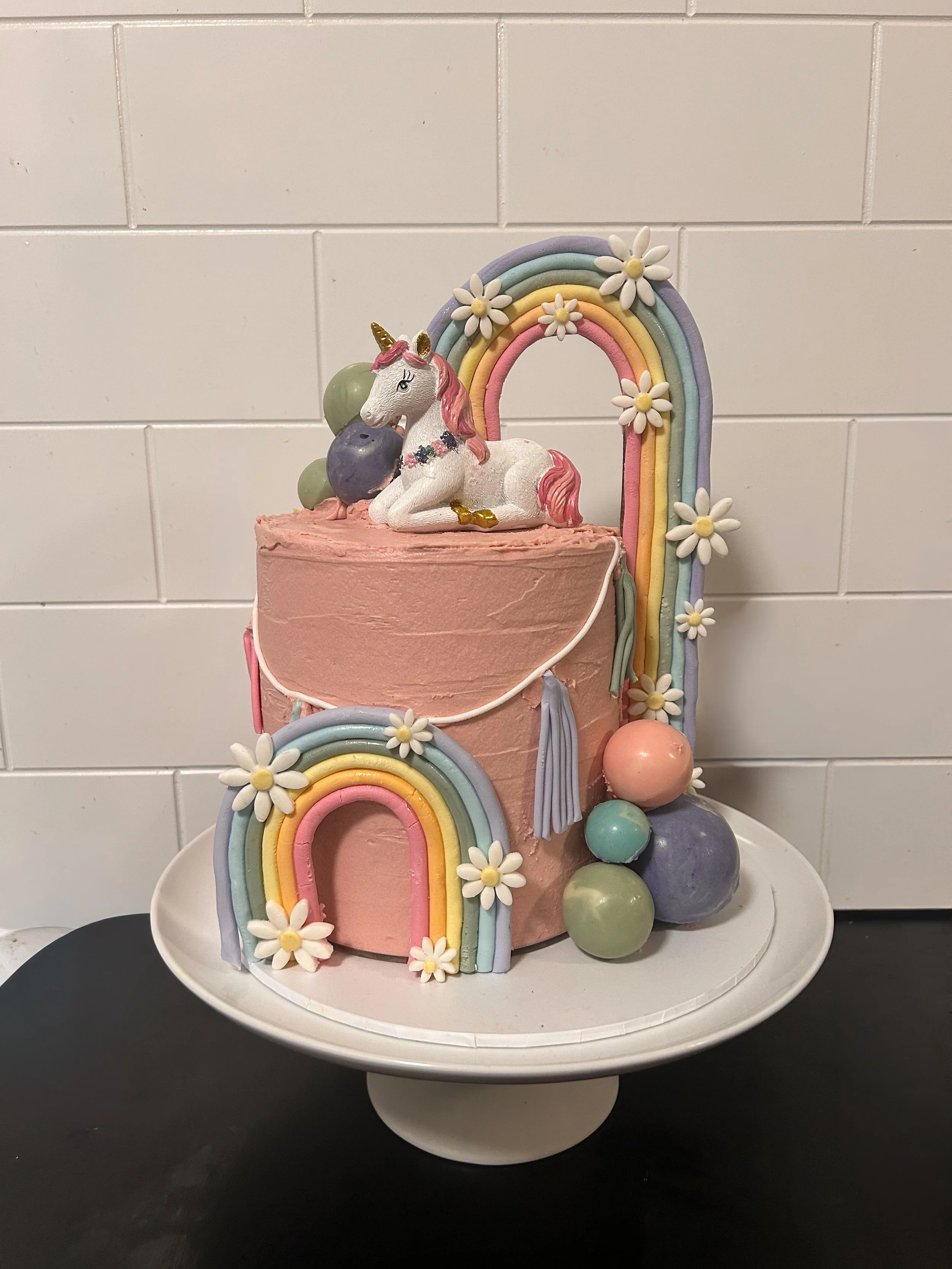 A pink unicorn-themed cake with colorful rainbow decorations and pastel-colored balls on a white cake stand.