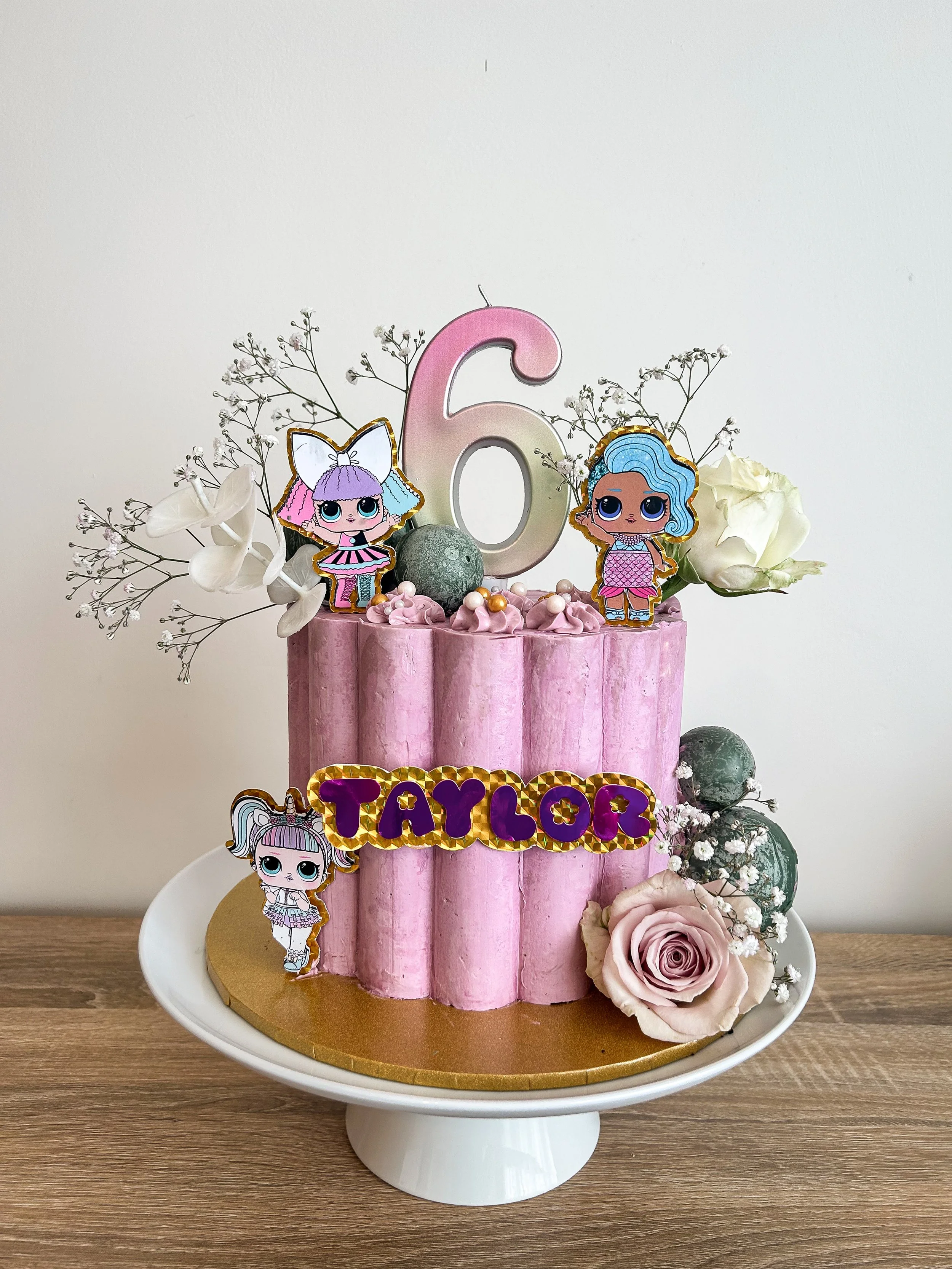 Pink birthday cake decorated with white flowers, a large pink number 6 candle, cartoon character toppers, and a gold and purple name plaque reading 'TAYLOR.' The cake is on a white cake stand on a wooden surface with a plain white background.