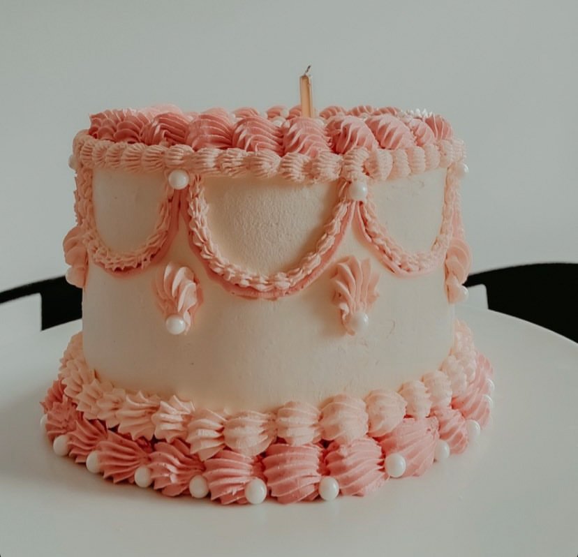 A round birthday cake decorated with pink and white icing, pearl-like embellishments, and pink icing garlands, with a single unlit candle on top.