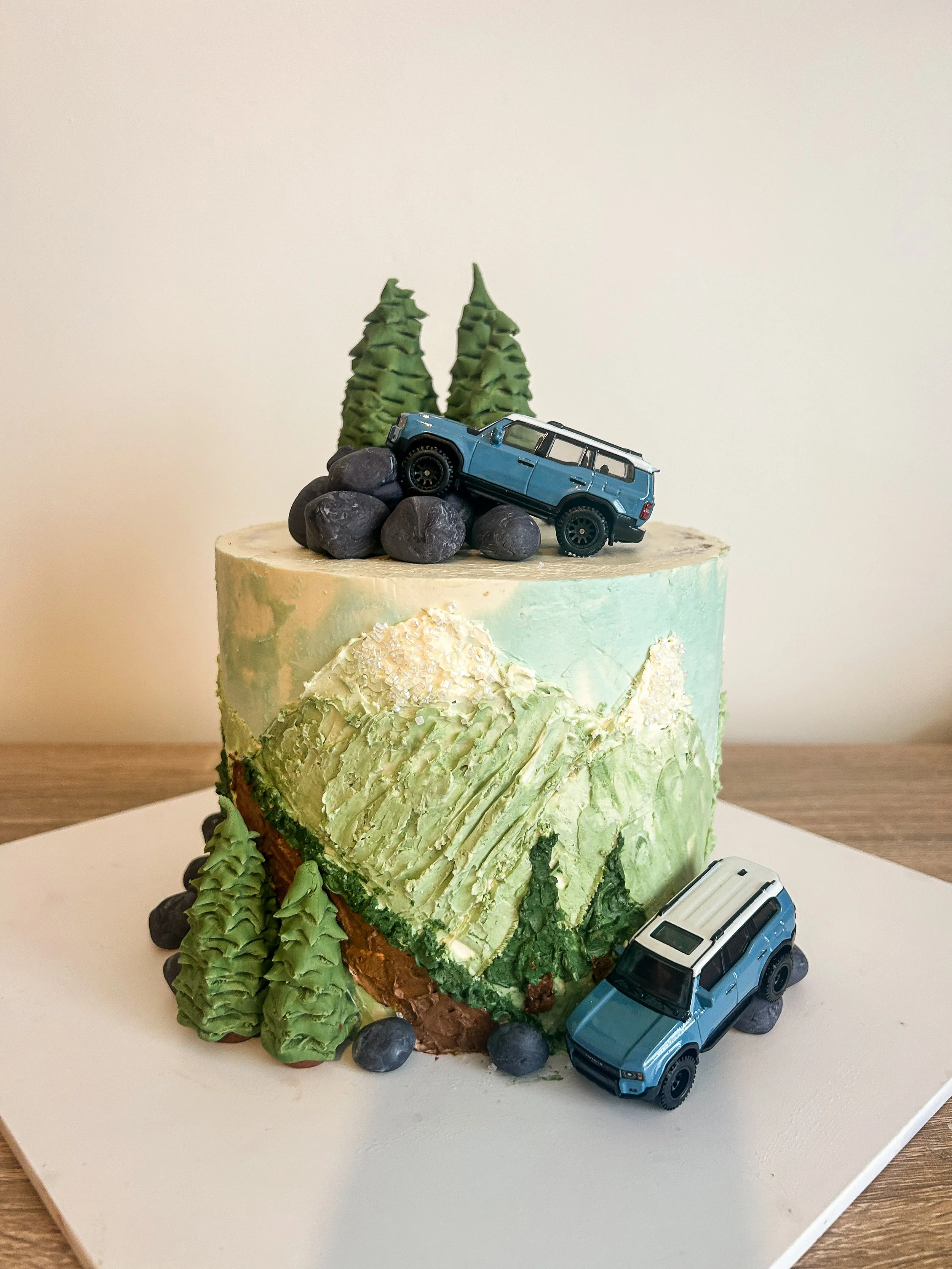 A two-tiered cake decorated with mountain, forest, and vehicle themes. The top tier features a miniature blue SUV model on rocks with forest trees behind it. The bottom tier displays a painted mountain landscape with trees and rocks at the base.