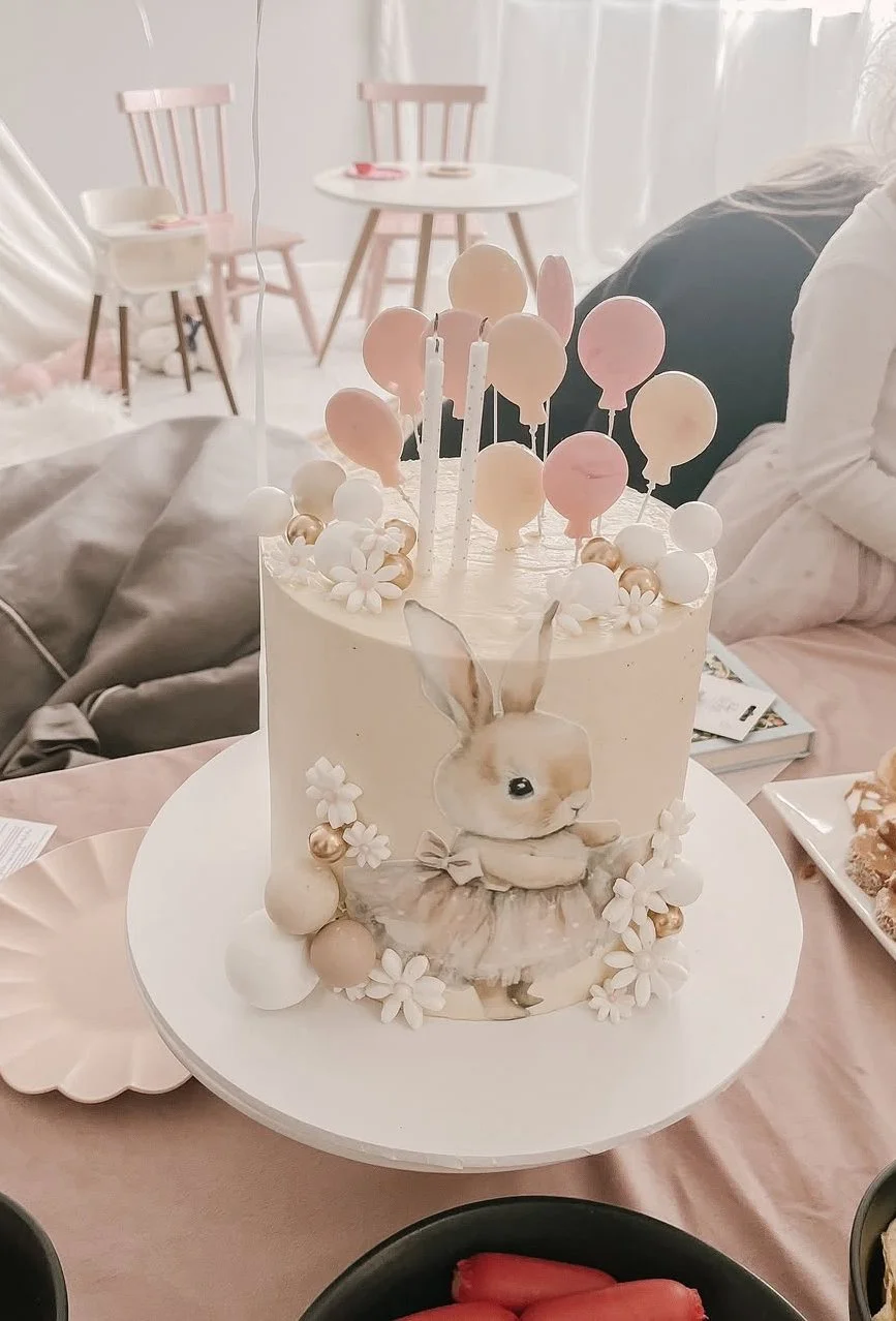 Decorative birthday cake with a bunny design, pink and white balloons, flowers, and candles on top, situated on a table with birthday decorations.