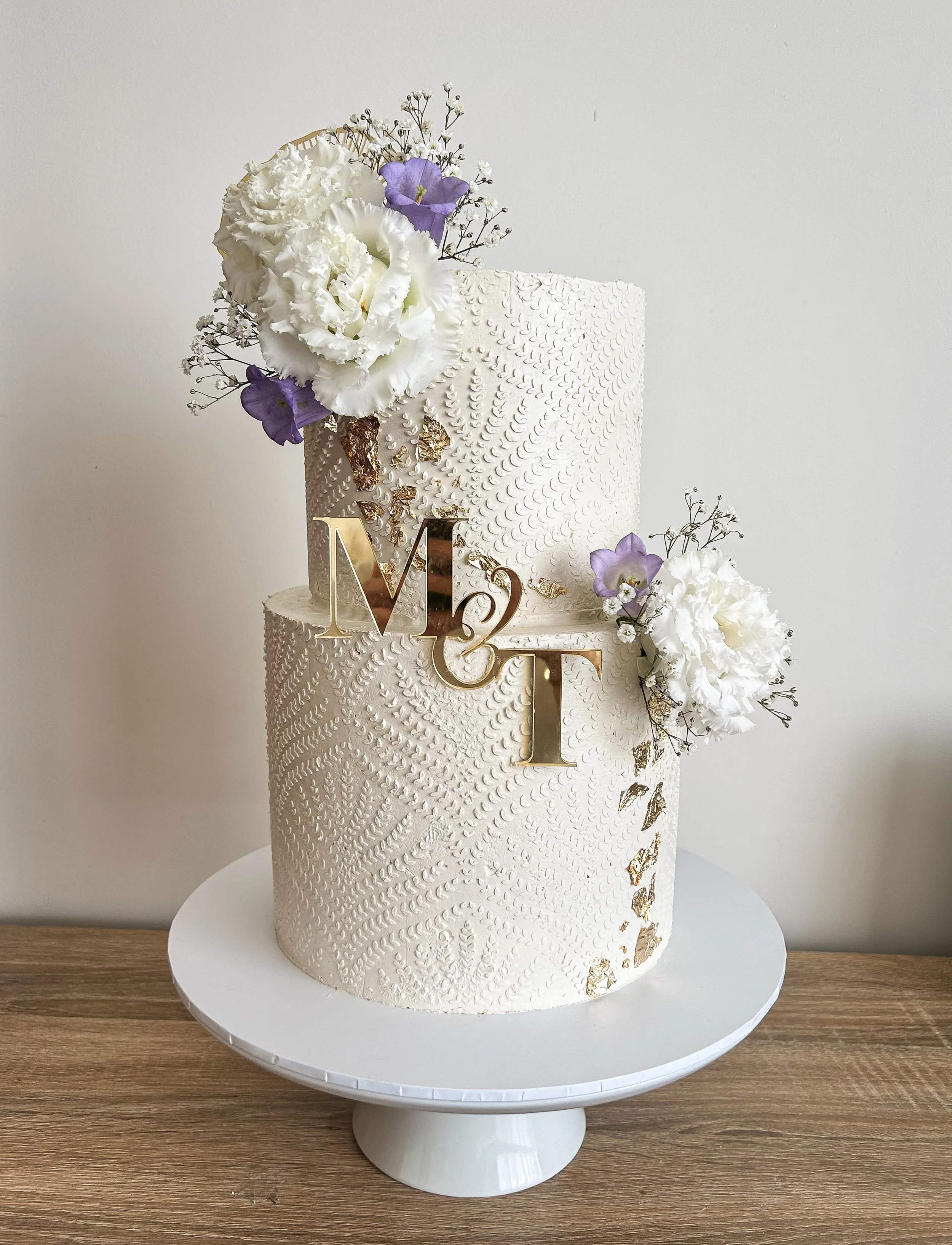 Two-tier white wedding cake with textured icing, decorated with white and purple flowers, gold butterfly accents, and a gold 'Mr & Mrs' cake topper, on a white cake stand.