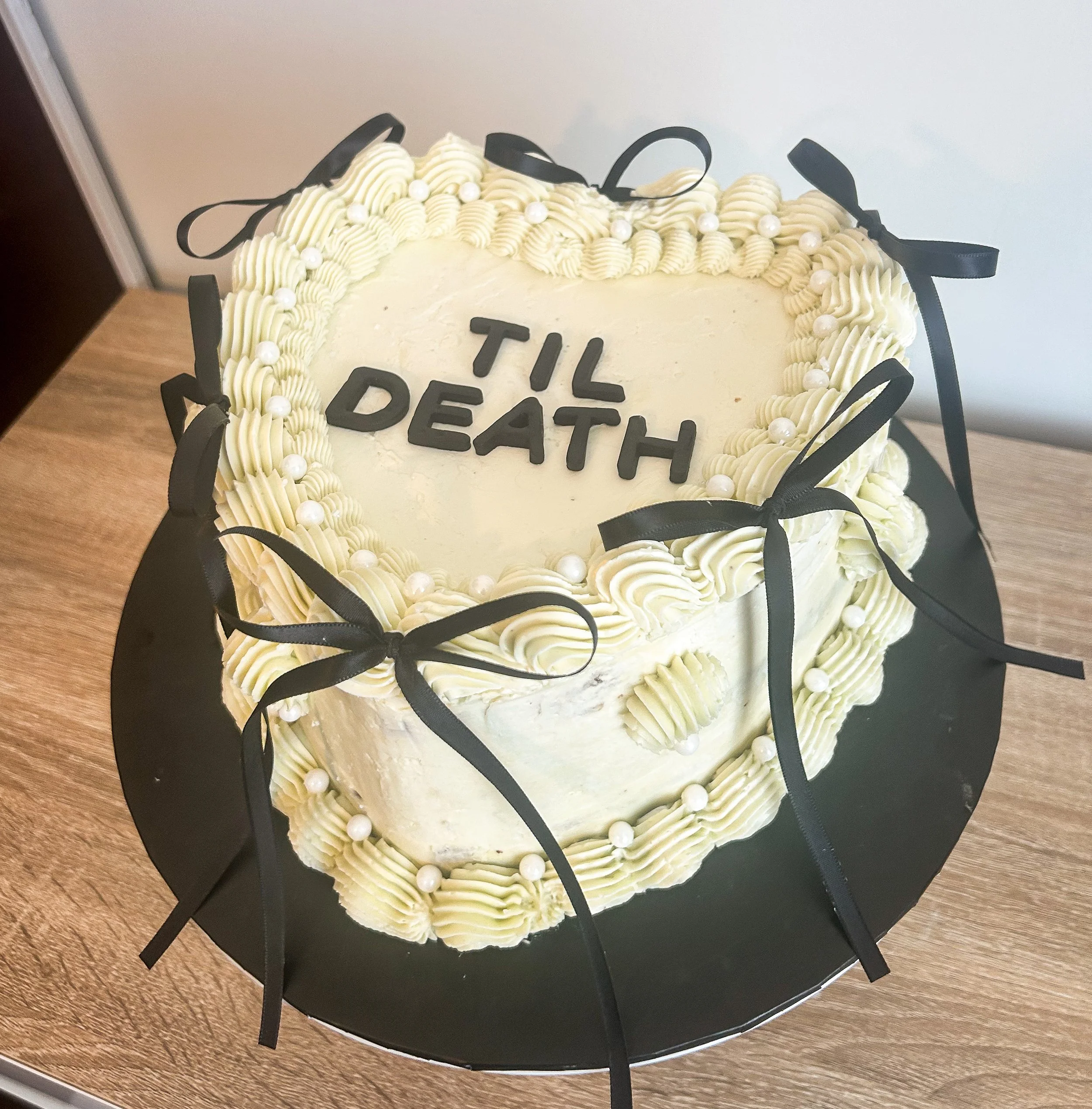 Heart-shaped cake with white frosting, black ribbons, white pearls, and the words 'TIL DEATH' written in black on top.