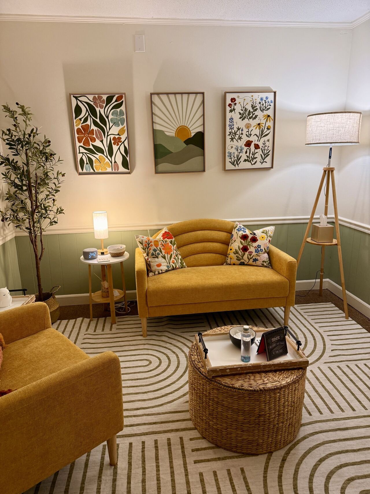A cozy living room with a yellow loveseat, a matching chair, and a wicker coffee table with decorative items. There are two standing lamps, a small side table with a lamp, and framed floral and landscape artwork on the wall. A striped rug covers the 