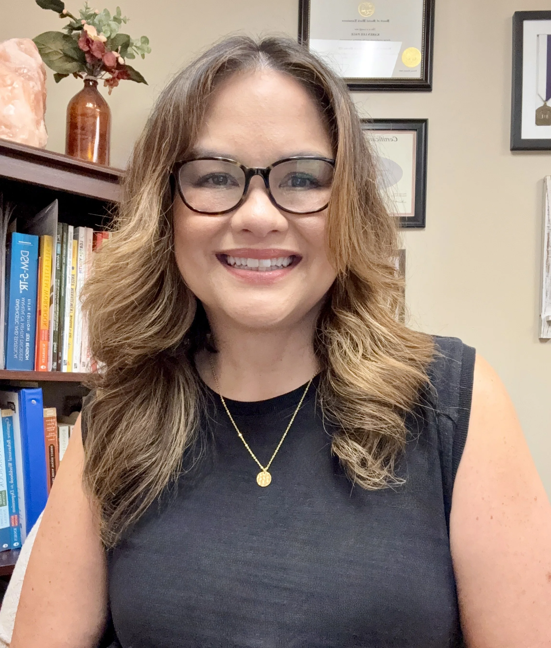 Smiling woman with wavy brown hair, wearing glasses, a black sleeveless top, and a gold necklace, sitting in a room with a bookshelf filled with books and framed certificates on the wall behind her.