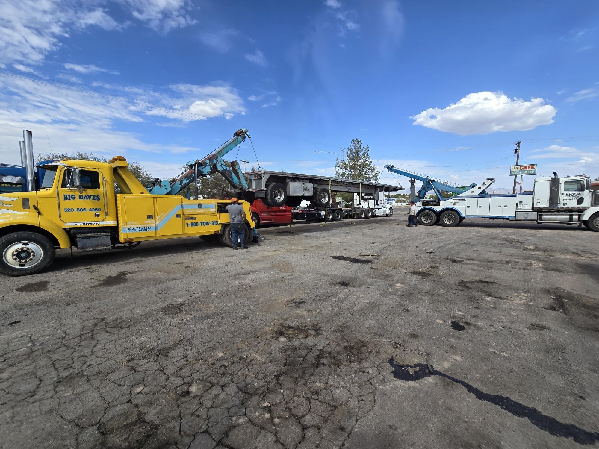 Two tow trucks are lifting and transporting vehicles in a lot with a cracked asphalt surface. The yellow tow truck to the left is lifting a small red truck and trailer, while the white tow truck to the right is nearby. There are a few people working around the trucks, and a sign indicating the presence of a cafe in the background under a partly cloudy blue sky.