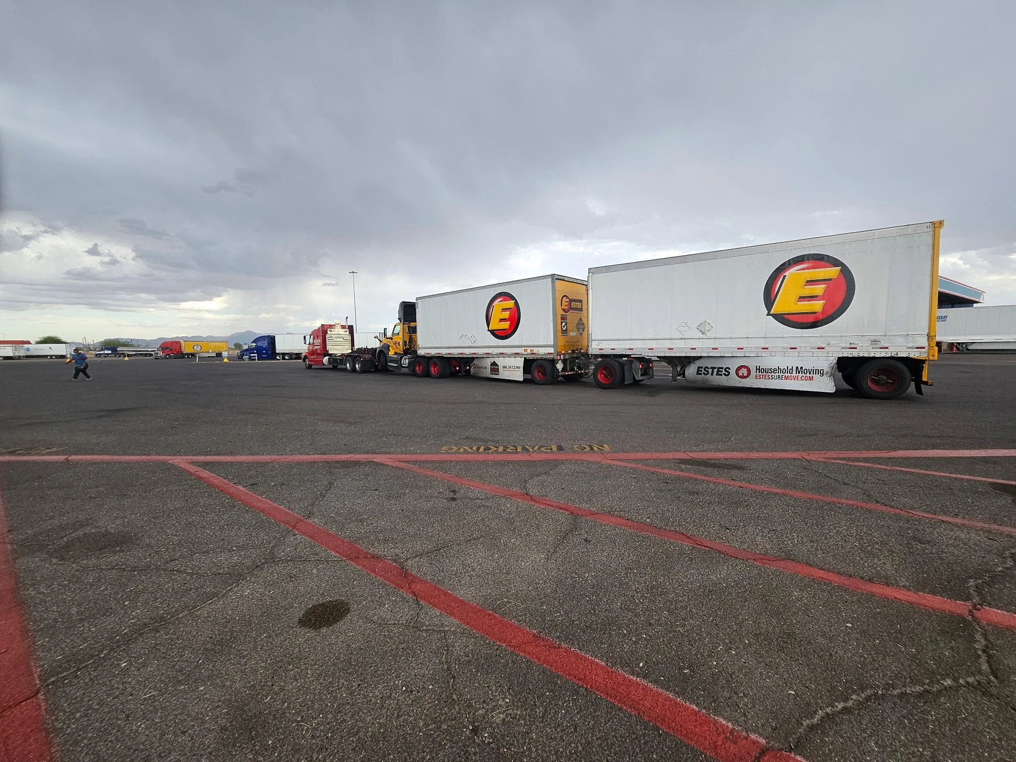 Large parking lot with several semi-trucks lined up, pilots walking, and a cloudy sky.