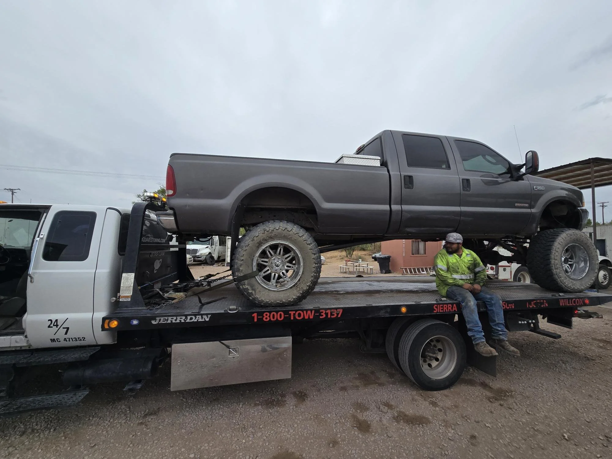 A tow truck with a gray pickup truck loaded on its bed, parked outdoors under a cloudy sky. A man in a green jacket and gray cap is sitting on the side of the tow truck.