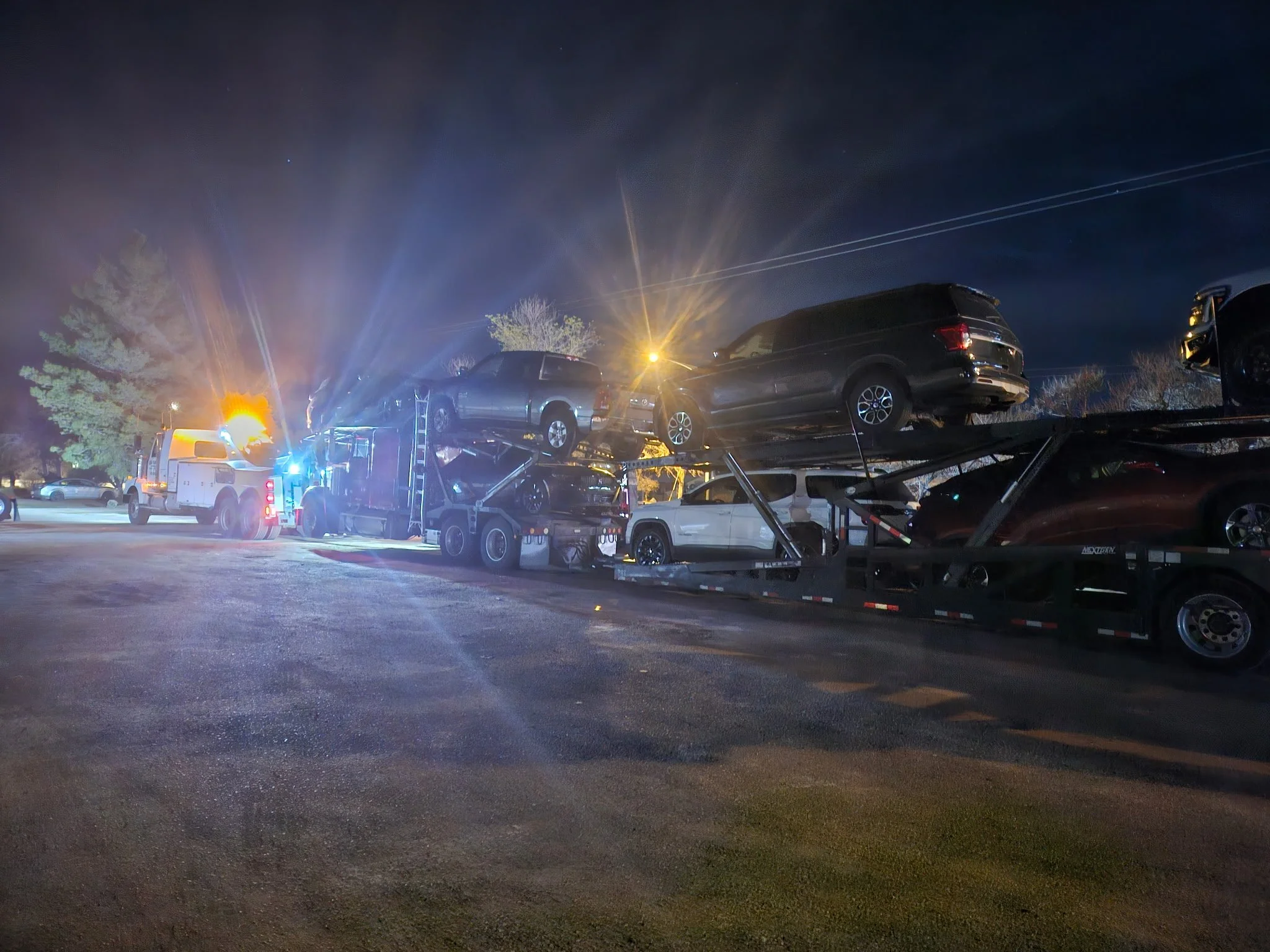 A car carrier truck at night transporting multiple black SUVs, with emergency lights flashing.