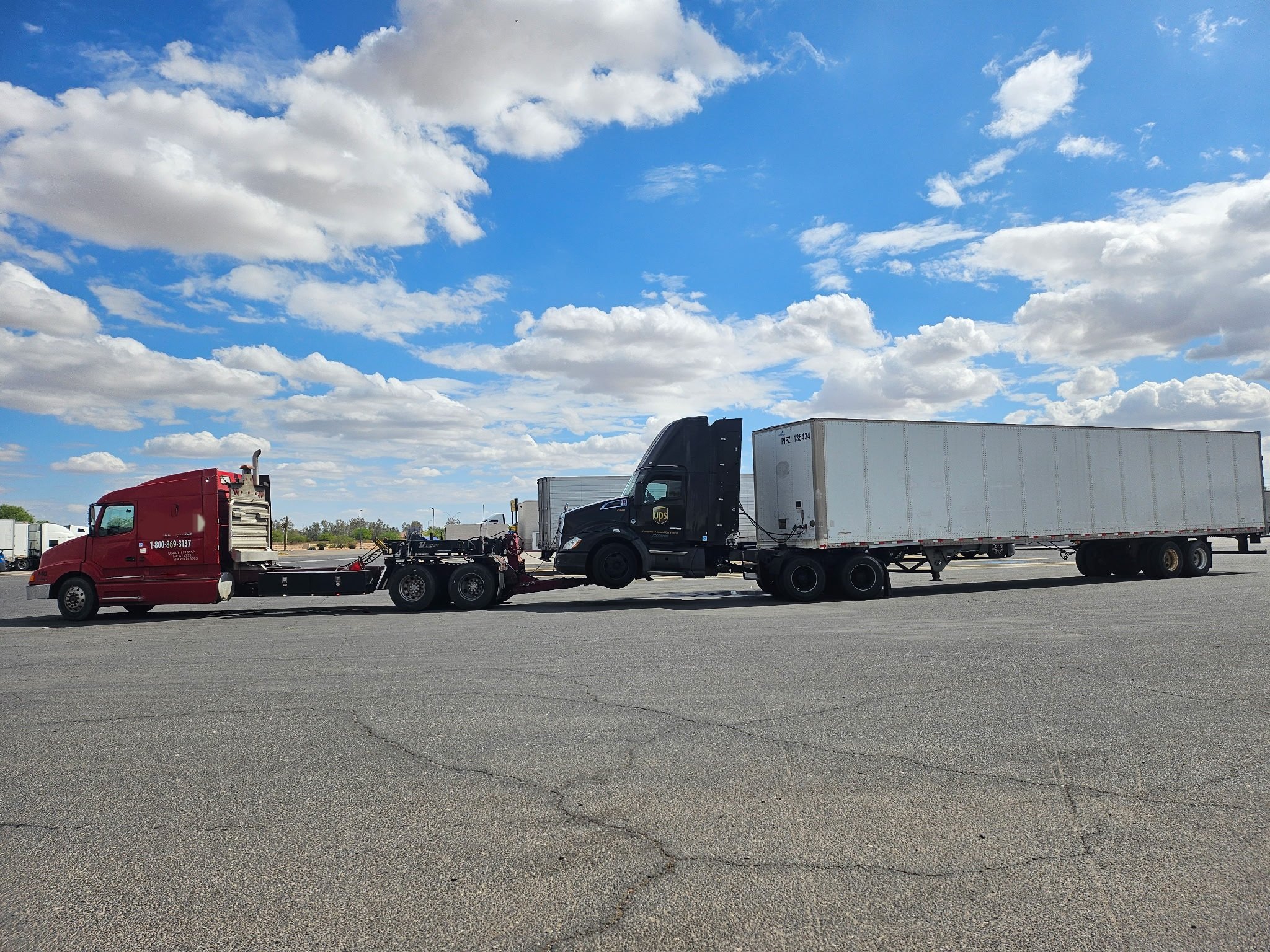 A semi-truck with a black cab and white trailer is parked on an empty lot under a partly cloudy sky.