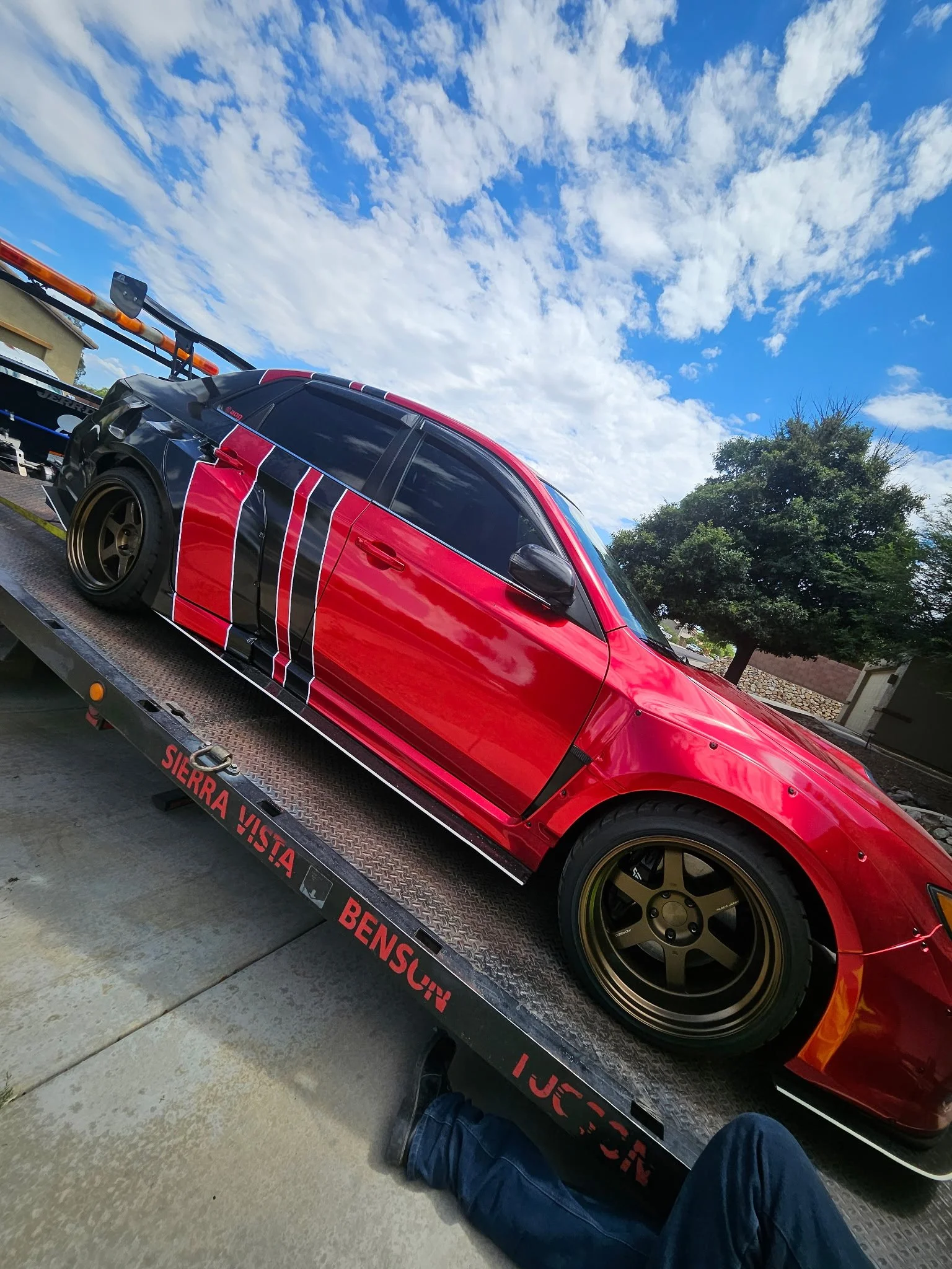 Red and black modified sports car with gold wheels on a flatbed trailer, outdoors on a sunny day with blue sky and clouds, trees in the background.