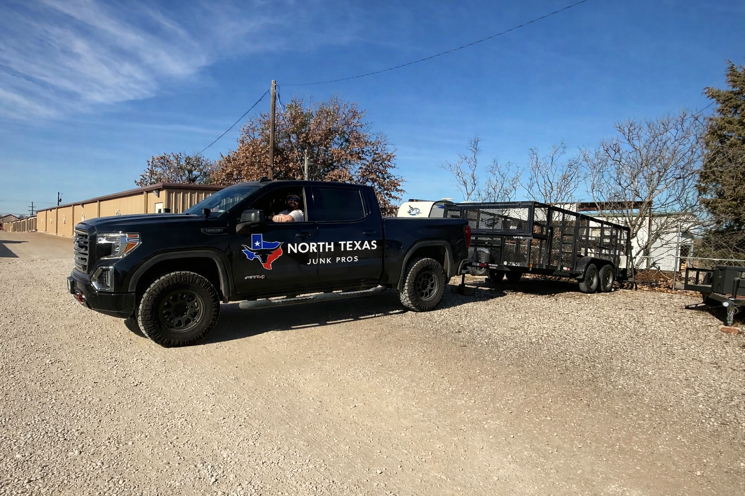 A black pickup truck labeled North Texas Junk Pros pulling a flatbed trailer, with a man sitting in the driver's seat, parked on a gravel lot under a clear blue sky.