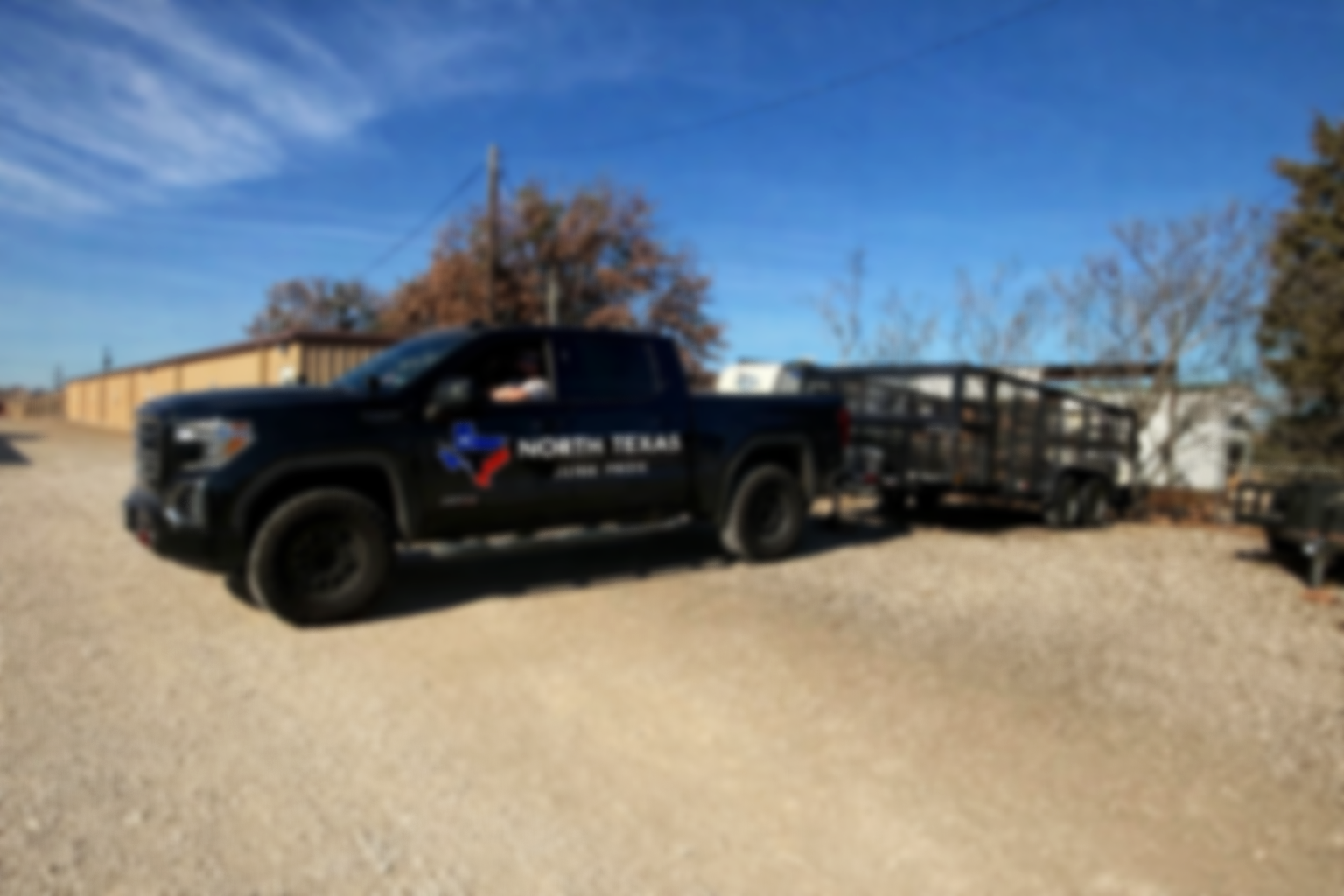 A black pickup truck with a North Texas logo on the door, attached to a trailer, parked on a gravel lot under a blue sky with a few clouds, with trees and a building in the background.