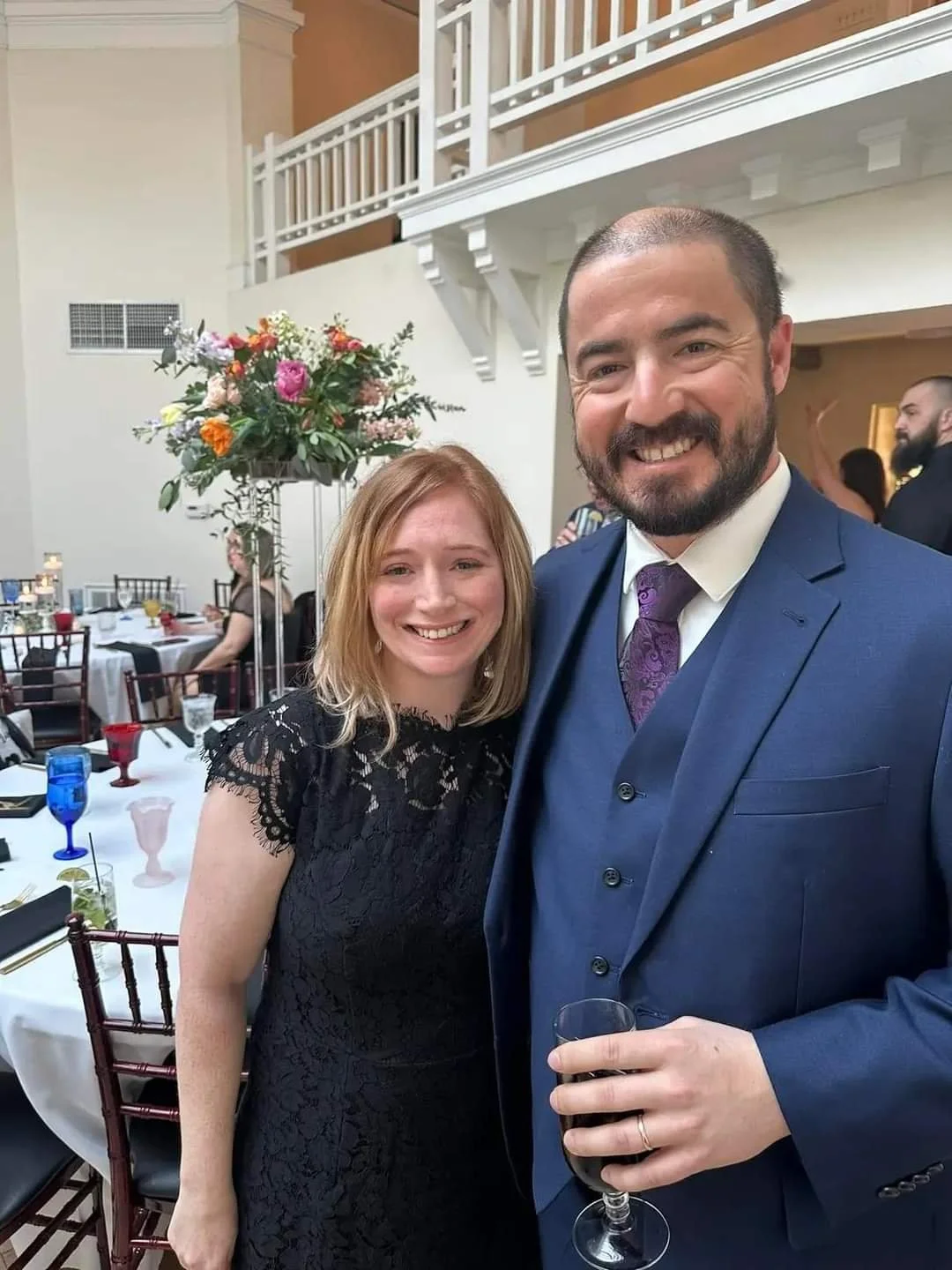 A smiling woman with blond hair wearing a black lace dress standing next to a smiling man with a beard dressed in a blue suit and purple tie holding a glass of red wine at a formal event or wedding reception.