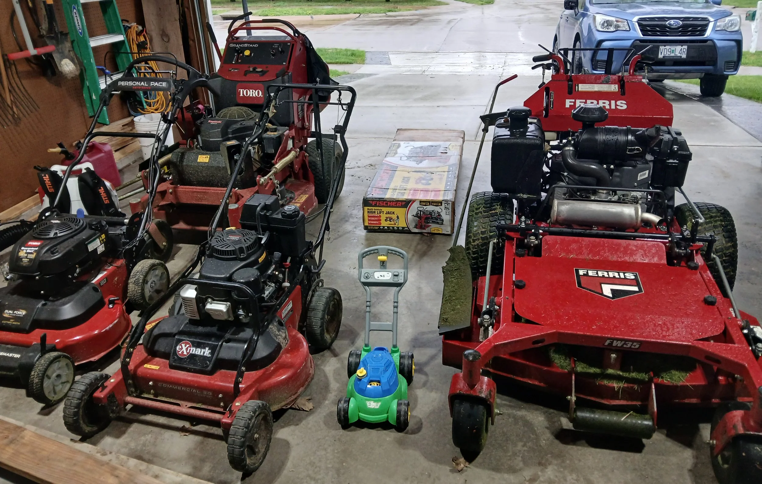 A garage with various lawn mowers, a small toy lawn mower, and two riding lawn mowers, with a car parked outside in the driveway.