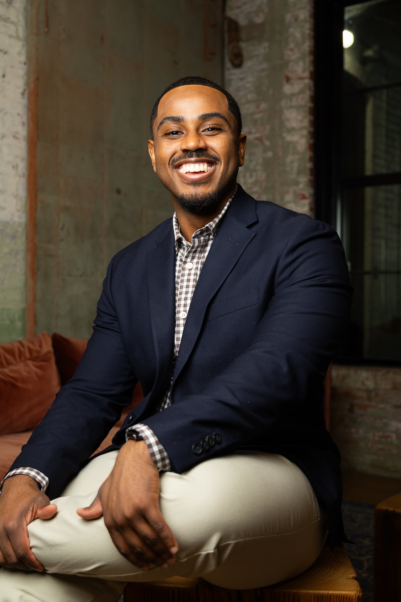 A smiling man wearing a navy blazer, checkered shirt, and beige pants sitting with one leg crossed over the other in front of a colorful, textured background with a window to his right.