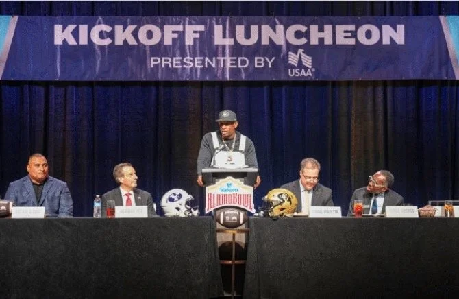 A panel of five men sitting at a long table on a stage with a purple banner that reads "Kickoff Luncheon Presented by USAA." One man is standing behind a podium, speaking. The stage has football helmets and a football on it, with one man wearing a football jersey and cap, and others dressed in formal attire.
