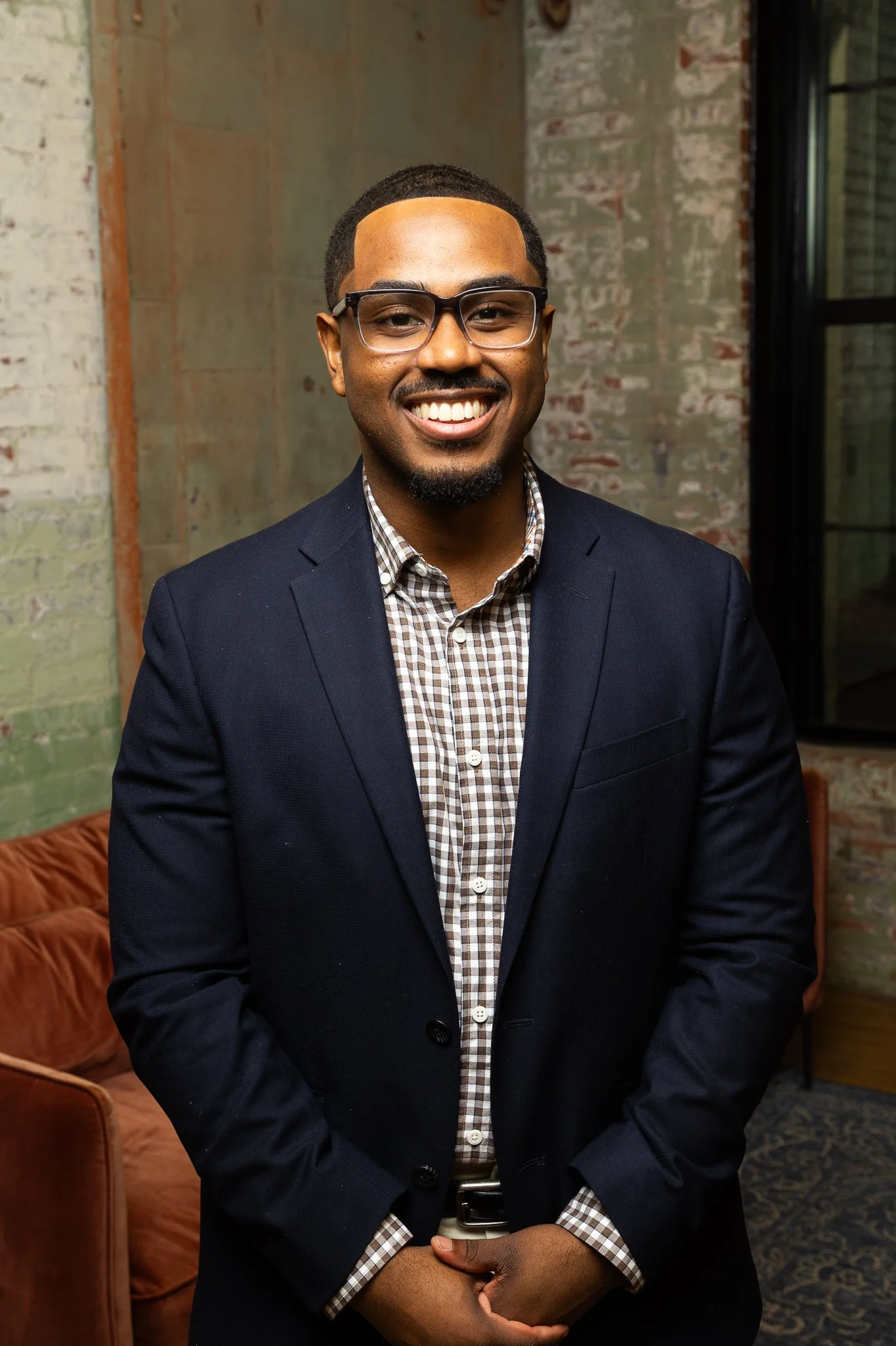 A smiling man wearing glasses, a checkered shirt, and a navy blazer standing indoors with exposed brick and a window in the background.