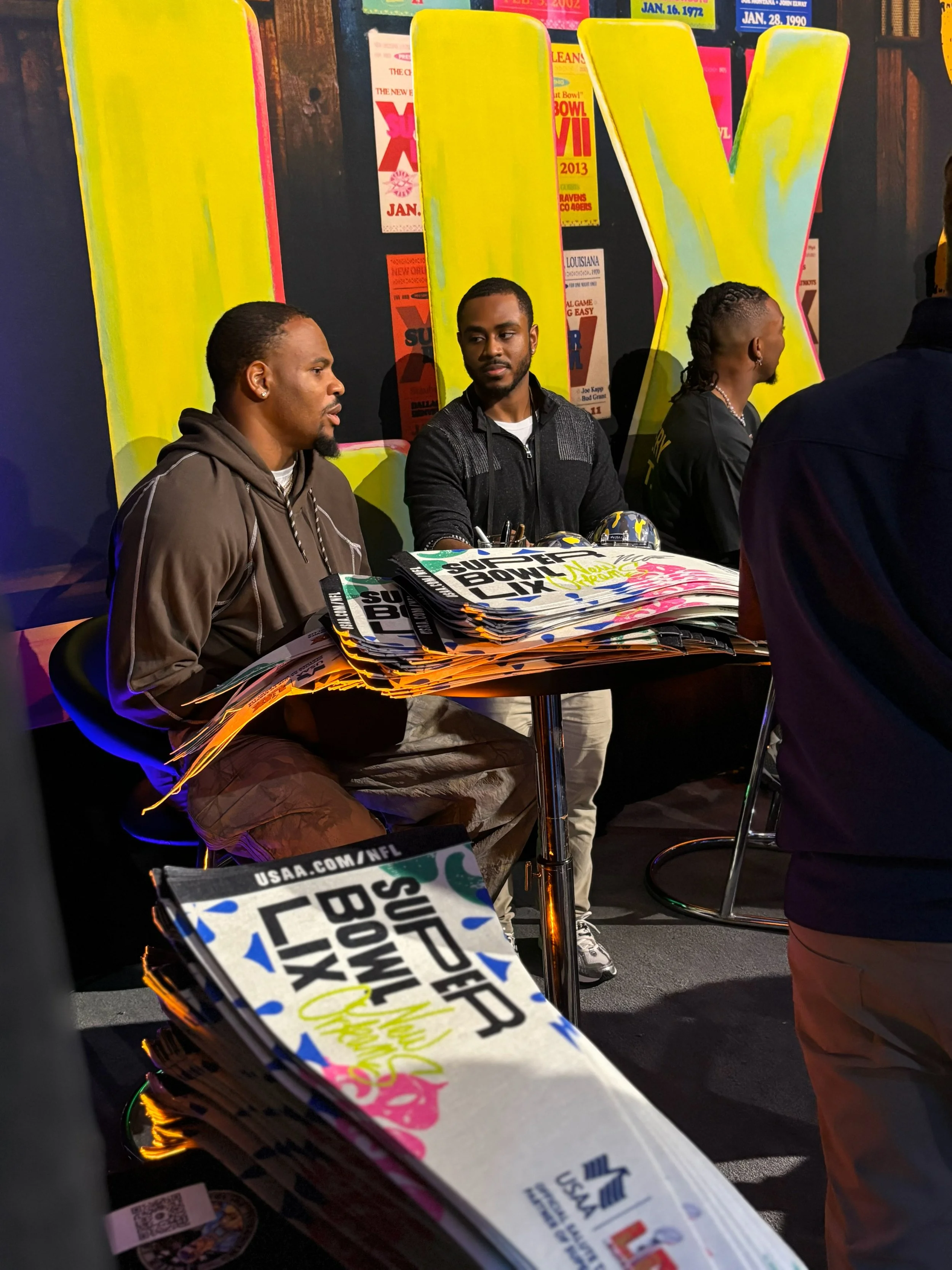 People sitting at a table during a Super Bowl LIX event, with large yellow letters in the background and stacks of Super Bowl LIX promotional materials on the table.