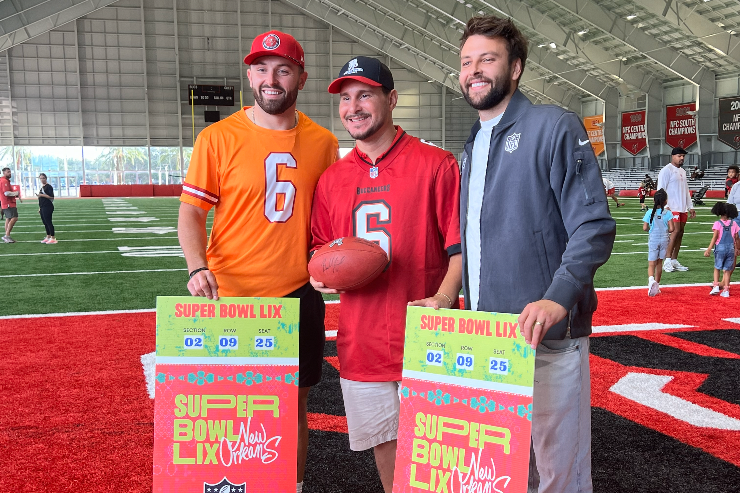 Three men holding Super Bowl LIX tickets inside an indoor football field. The man on the left is wearing an orange jersey with number 6 and a red cap. The man in the middle is wearing a red jersey with number 6, holding a football. The man on the right is wearing a gray jacket and holding a Super Bowl LIX ticket. In the background, children and other people are on the field with banners hanging from the ceiling.