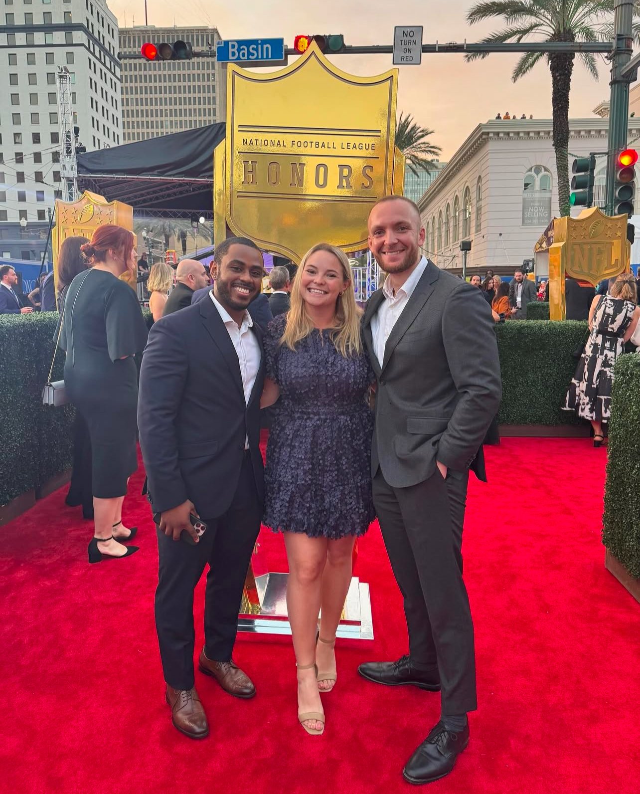 Three people, two men and one woman, standing on a red carpet at a formal event, with a large gold sign that says 'Honors' in the background during sunset.