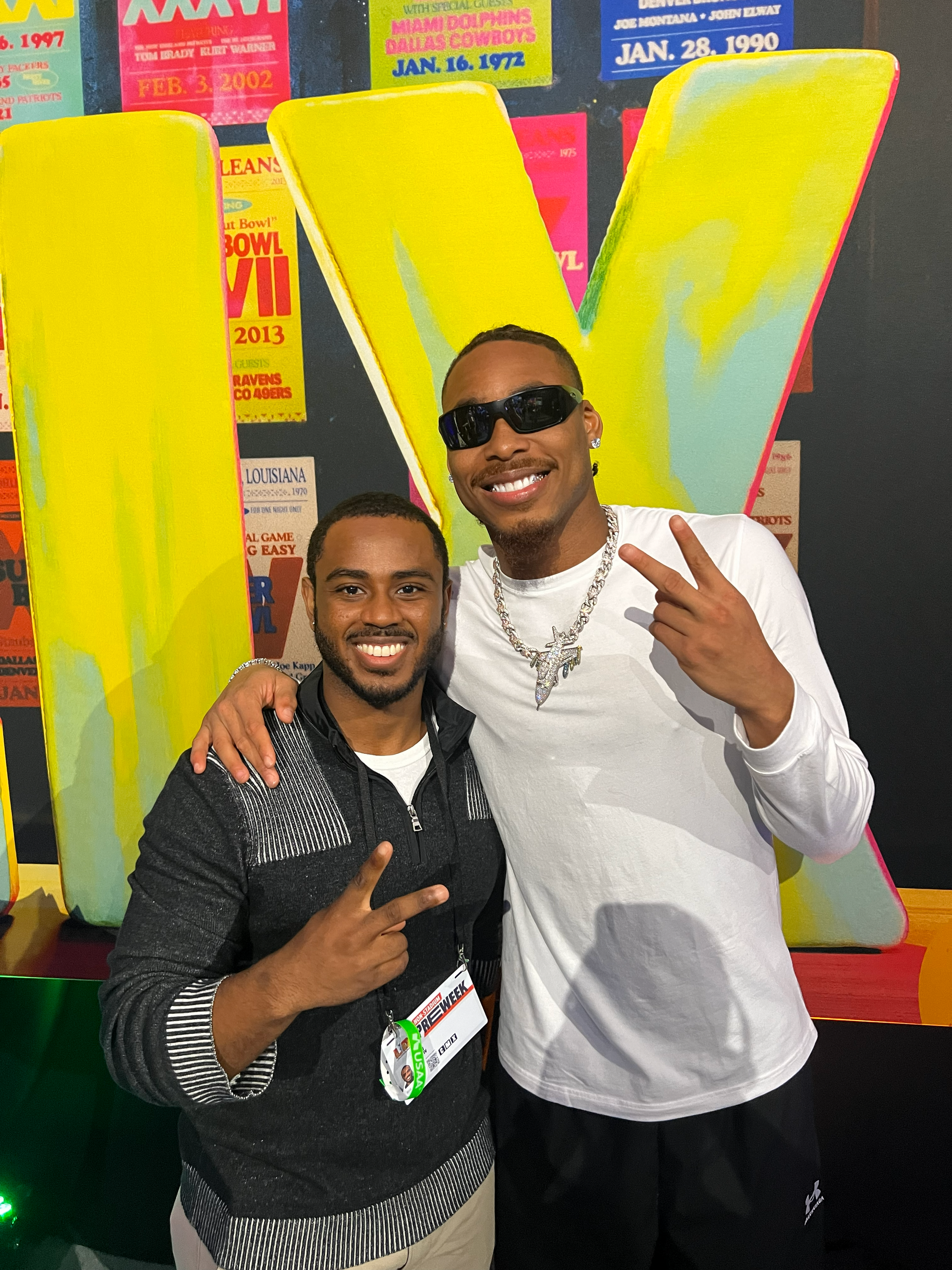 Two men smiling and making peace signs with their fingers, standing in front of a large yellow and teal 'V' letter display with colorful event posters in the background.