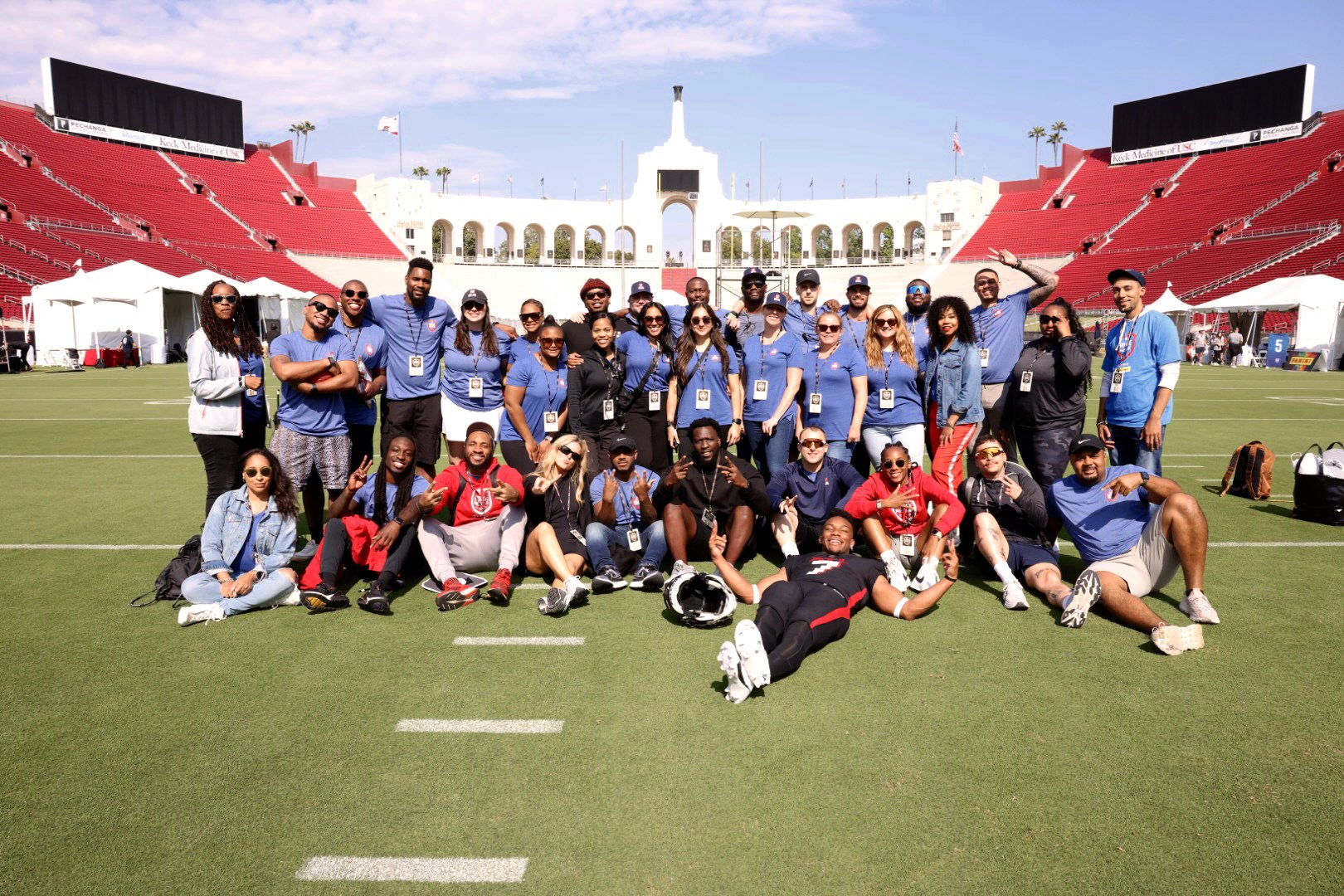 Group of around 30 people standing and sitting on a football field, posing for a photo, with a stadium structure and red bleachers in the background, under a partly cloudy sky.