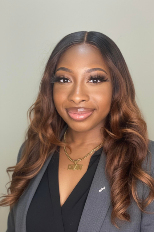A woman with long wavy brown hair, wearing makeup, a gray blazer, and a black top. She has a gold chain necklace and is smiling at the camera.
