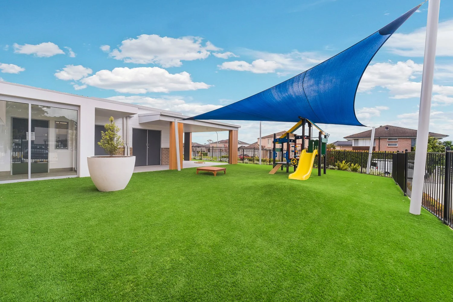 Children's playground with a yellow slide, climbing structure, blue shade sail, green artificial grass, and a modern building with large windows and a potted plant outside.
