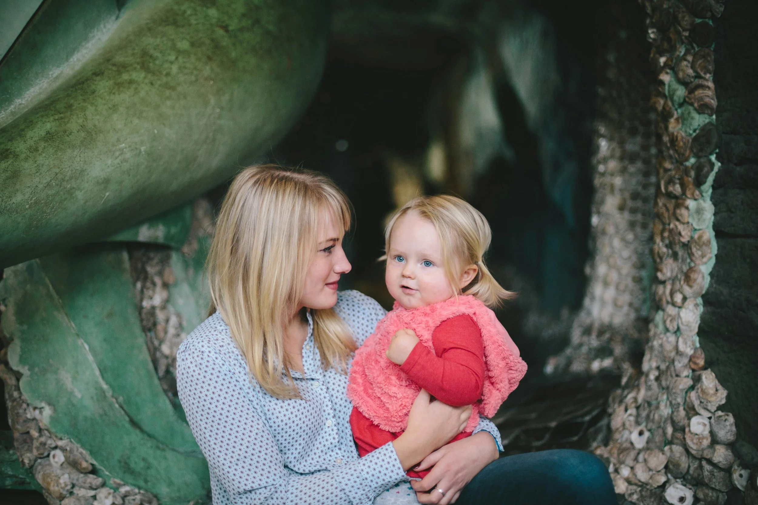 A woman with blonde hair holding a young girl with blonde hair and blue eyes, sitting together in a natural setting with large green rocks and a shell-covered surface behind them.