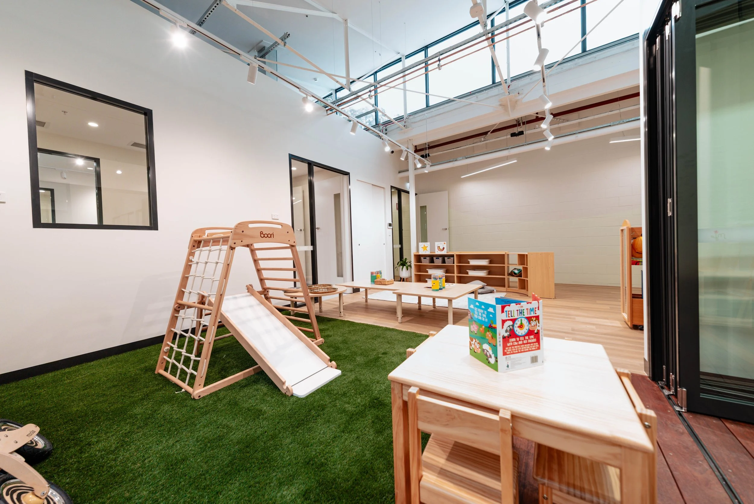 A modern indoor playroom for children with a small slide, low tables, shelves with toys, and a green artificial grass floor area, illuminated by natural light through large windows.