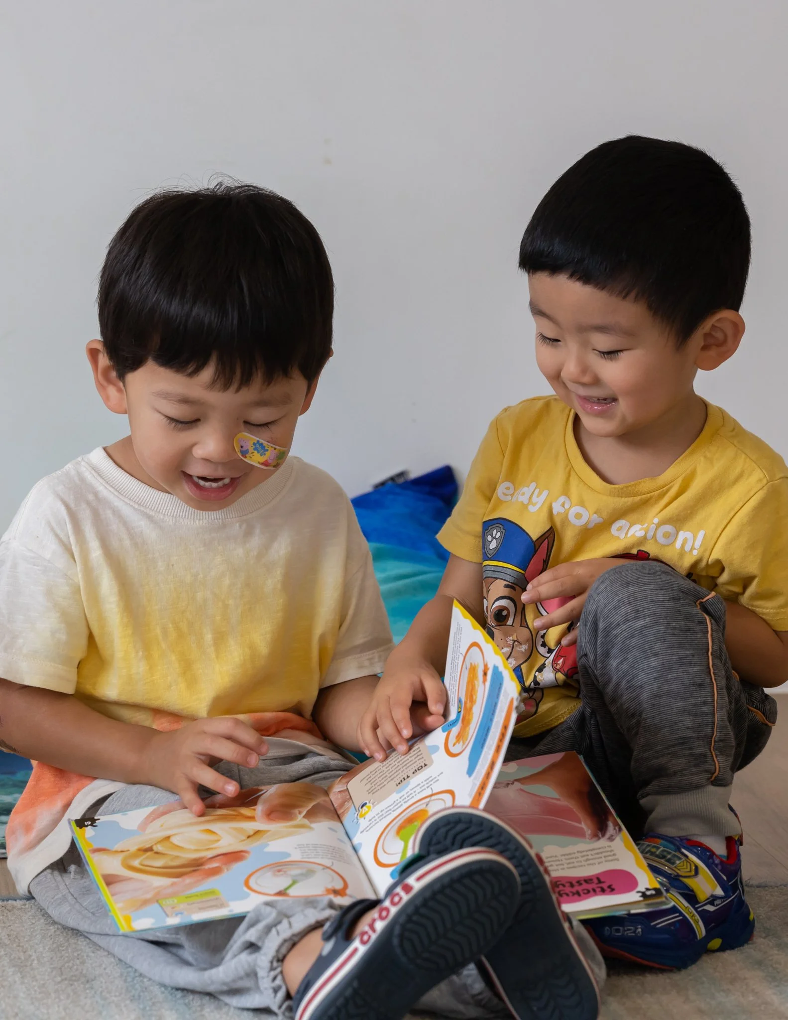 Two young boys sitting on the floor, reading a colorful book together, smiling and appearing engaged.