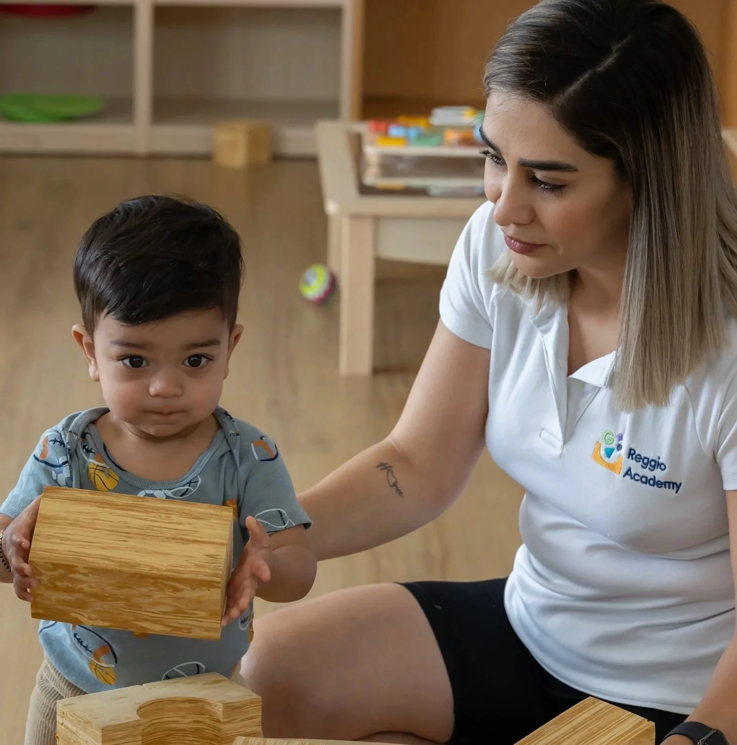 A woman and a young boy in a classroom, with the woman helping the boy hold a wooden block while other wooden blocks are on the table.