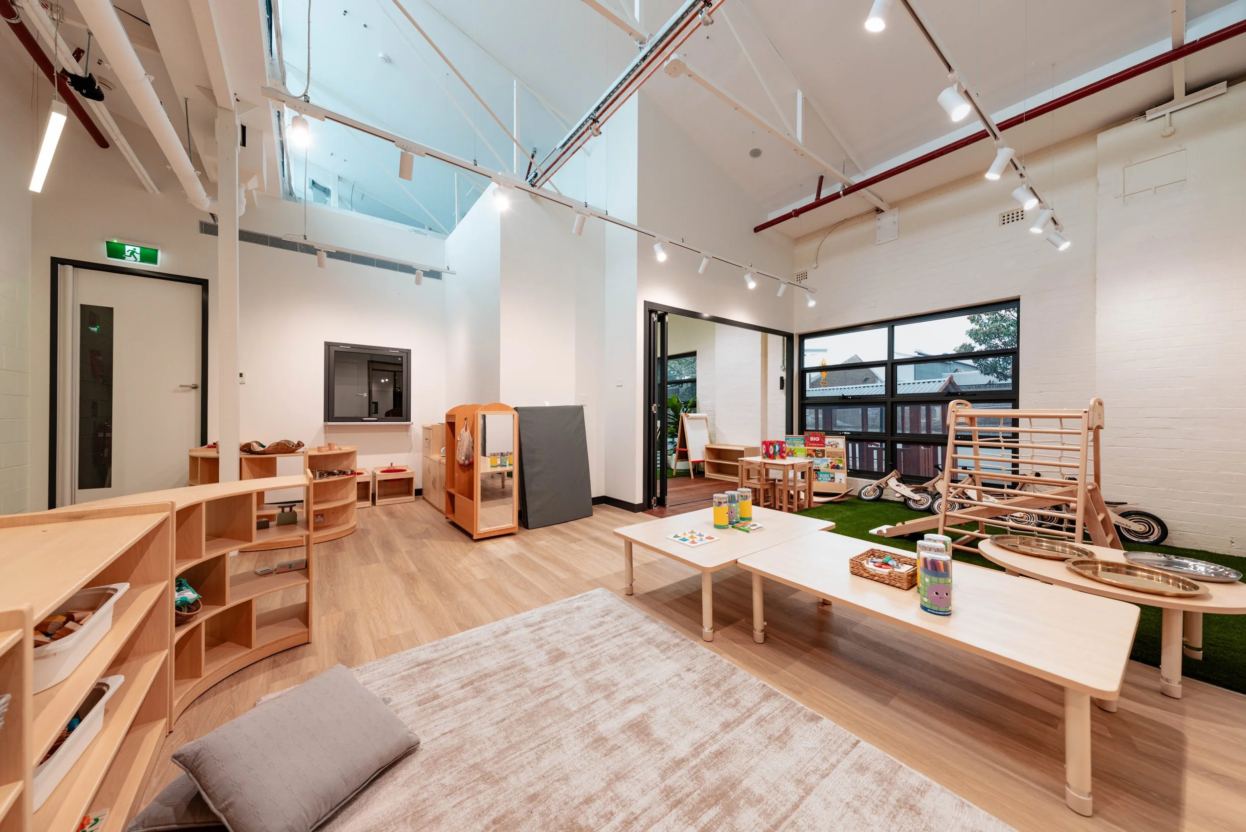 Bright, spacious indoor play area for children with wooden furniture, toys, a small grassy play zone with bikes, and large windows providing natural light.