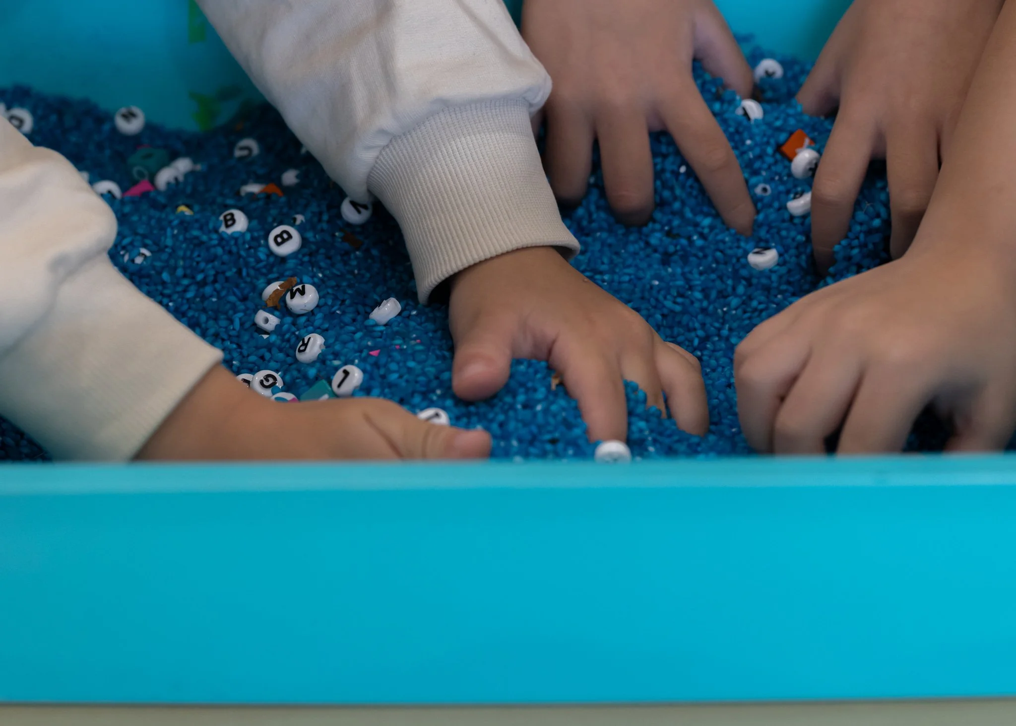 Children's hands playing in a blue sensory bin filled with small beads and lettered alphabet pieces.