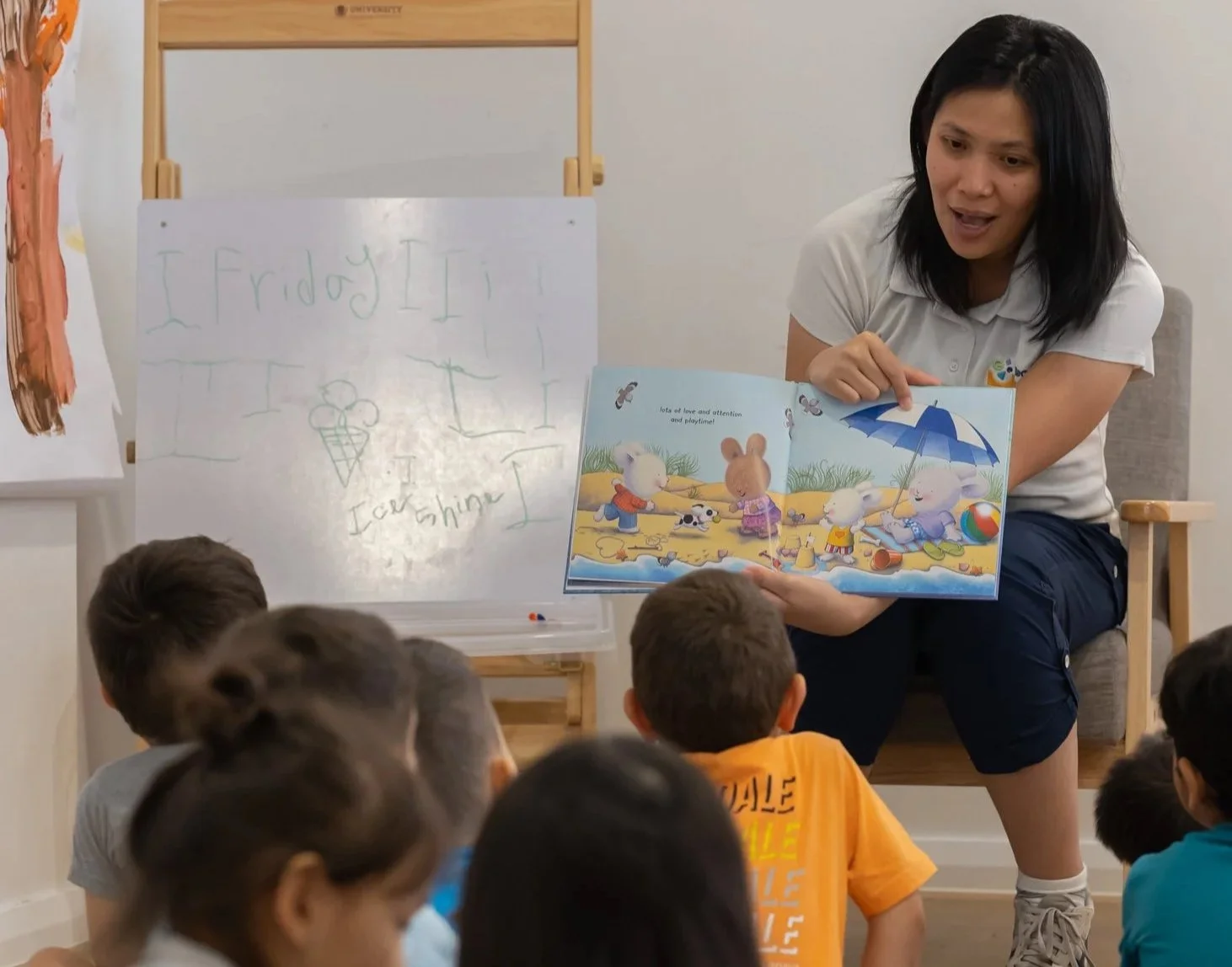 A woman is reading a colorful children's book to a group of children seated on the floor. The book features animals on a beach and has text about love and attention. A whiteboard behind her has handwritten words and a drawing of an ice cream cone.