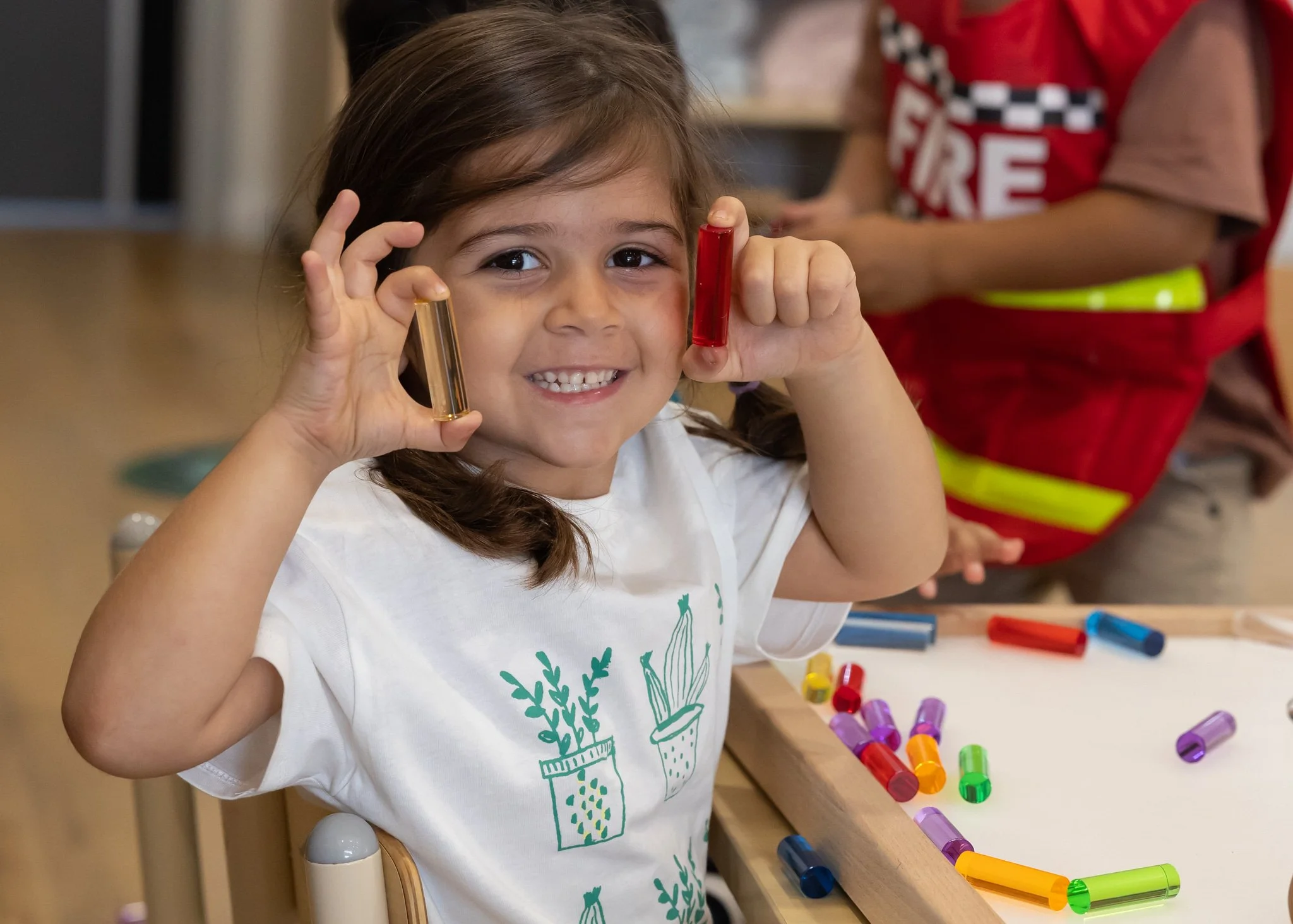 A young girl smiling and holding a small test tube with a gold-colored liquid in her left hand and a red test tube in her right hand, sitting at a table with colorful test tubes and supplies, in a classroom or activity setting.