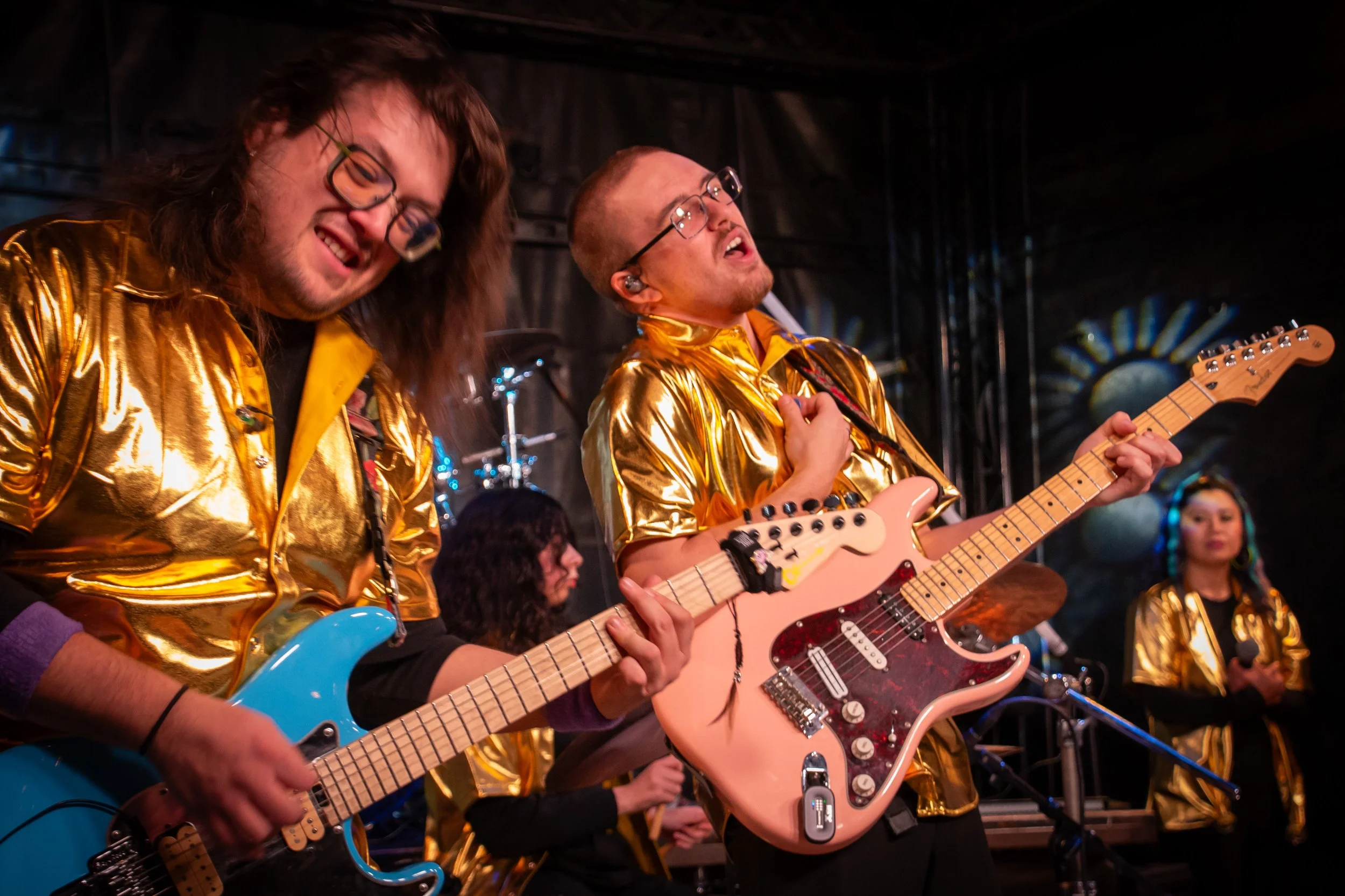 Musicians wearing shiny gold shirts performing on stage with guitars, one playing a blue electric guitar and another with a pink electric guitar, in a lively performance setting.