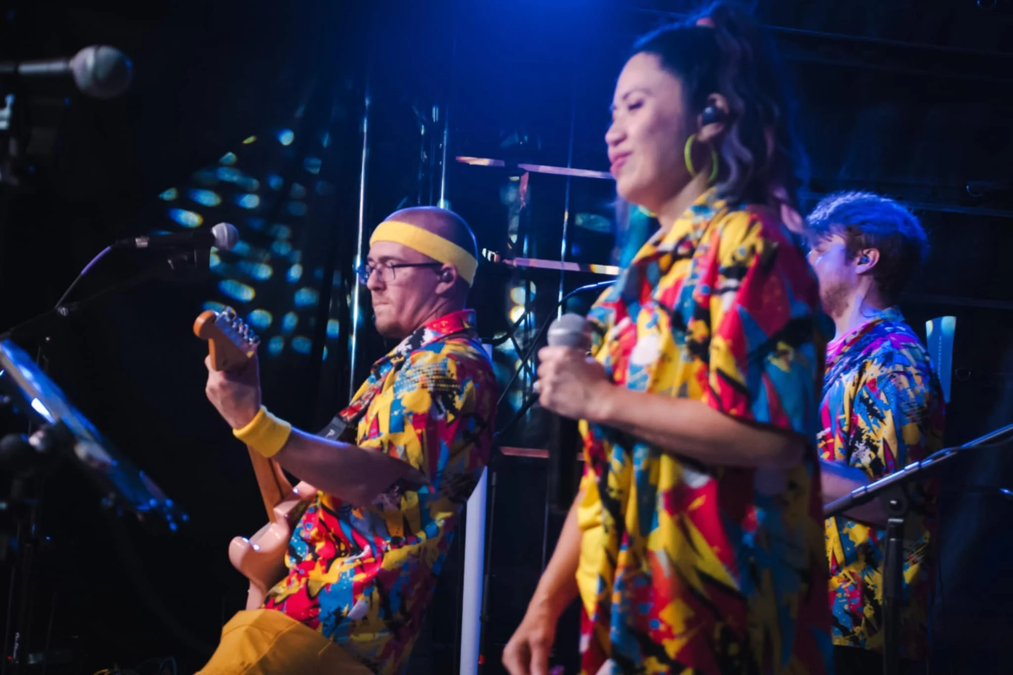 Band performing on stage with colorful outfits, one member playing guitar, others singing into microphones, stage lighting creating a vibrant atmosphere.