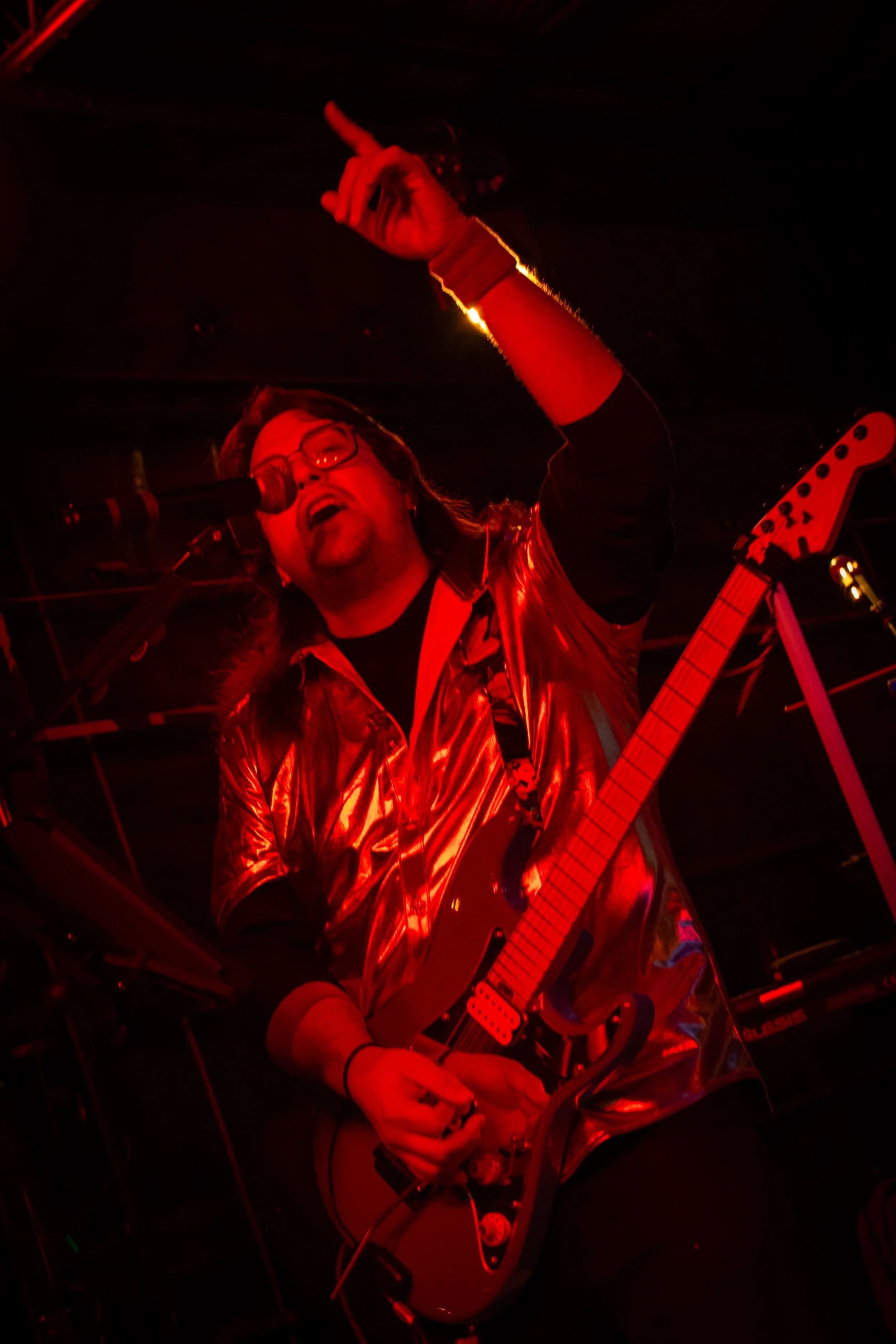 A guitarist wearing a shiny gold shirt and glasses, performing passionately on stage with a red lighting effect.