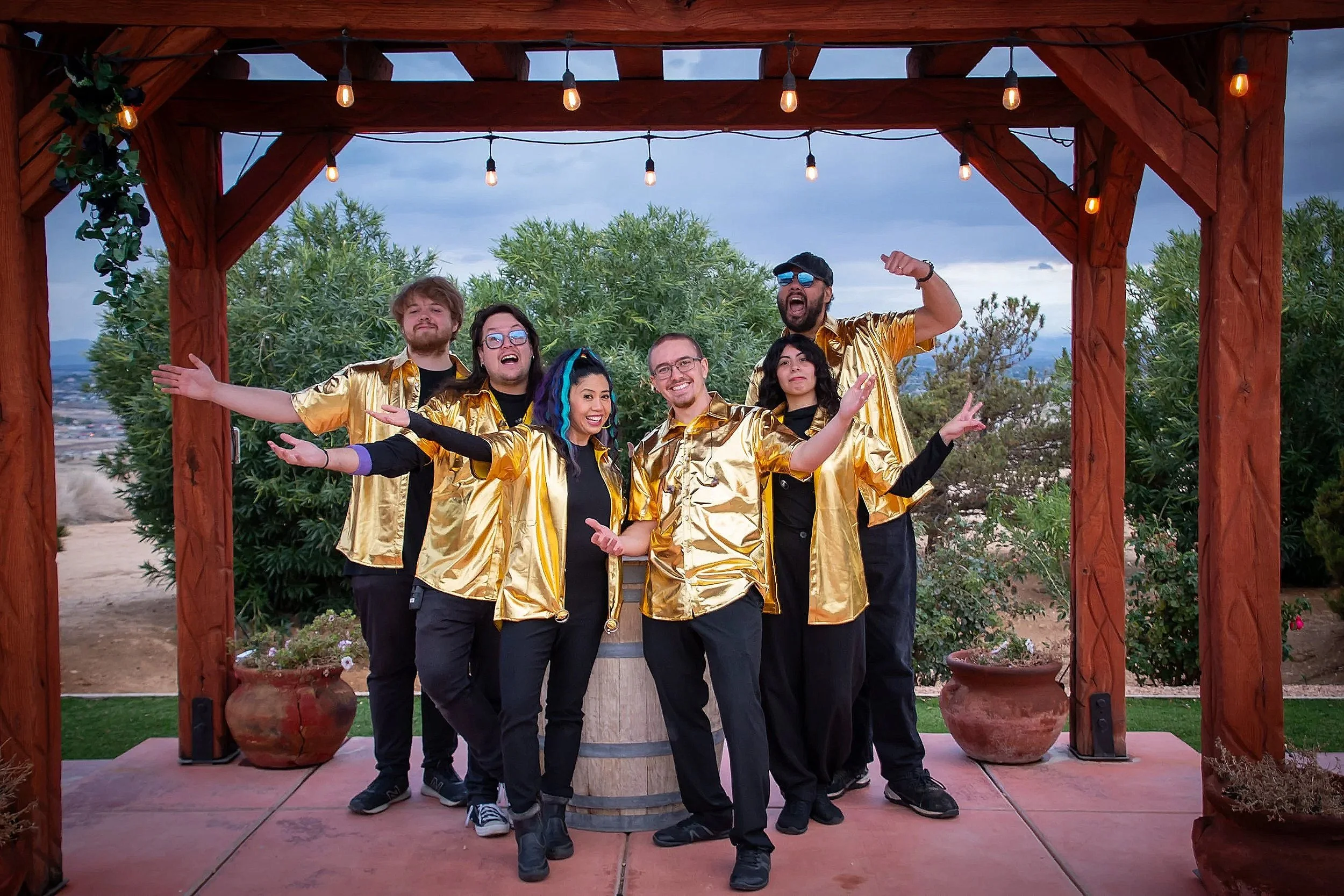 Group of six people posing outdoors under a wooden pergola, wearing matching golden shirts, with a scenic background of trees and a cloudy sky.