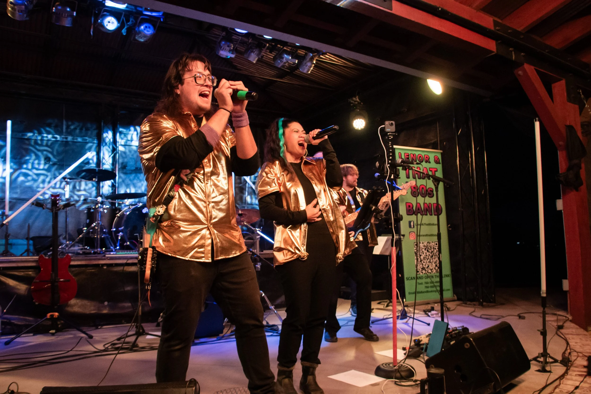 A band performing on stage with two singers wearing gold shirts, and a guitarist in the background, in a dimly lit venue.