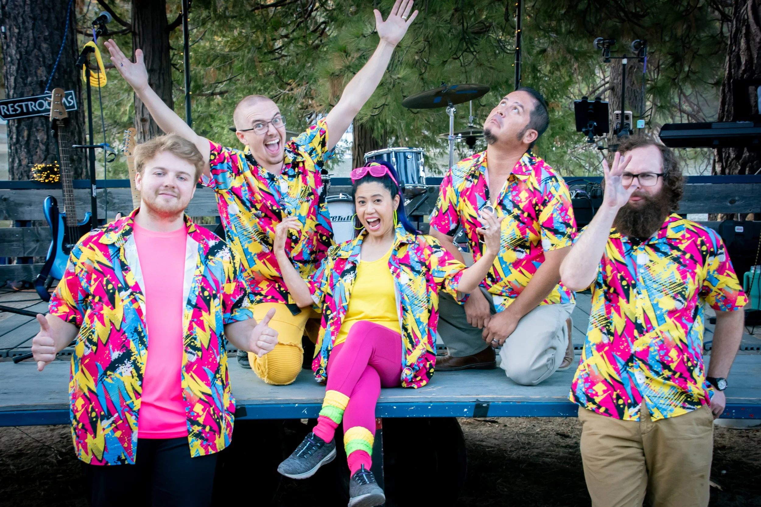 Group of five people wearing colorful 80s-style shirts, posing on a stage outdoors with musical instruments in the background.
